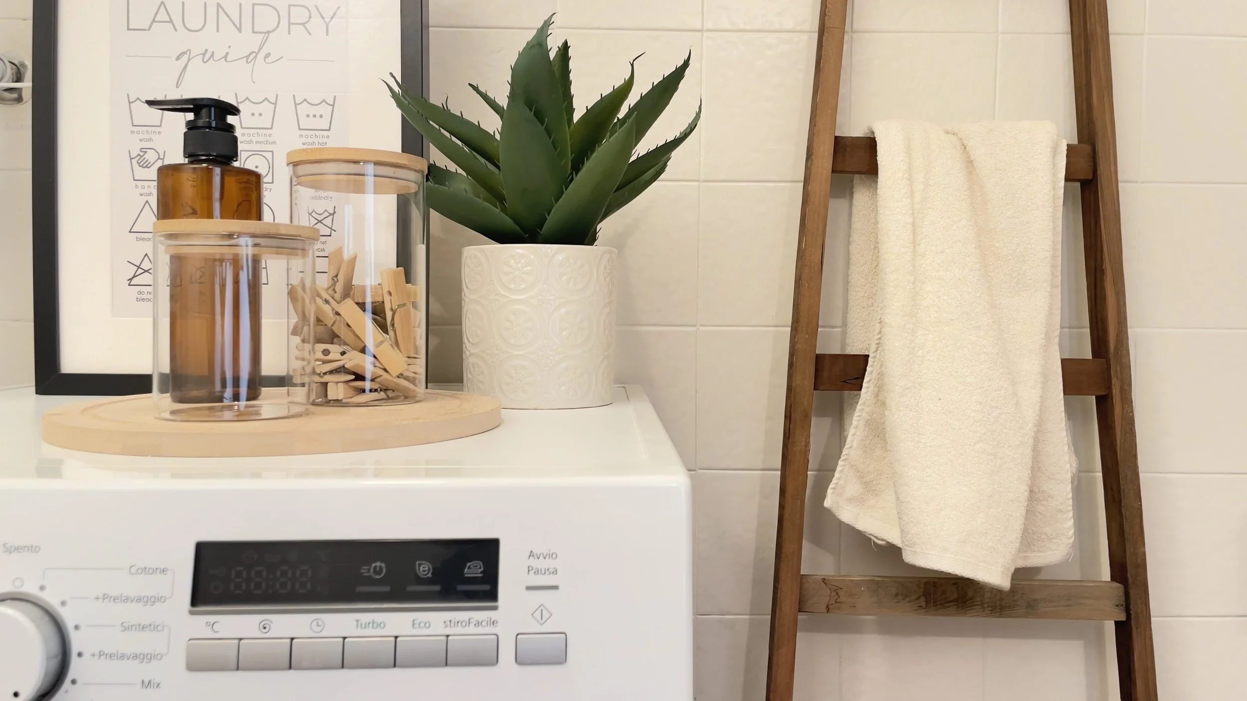 Lavatrice con contenitore di detersivo, mollette in un vaso trasparente, pianta in vaso bianco, scala in legno con asciugamano appeso, guida per il bucato su sfondo.