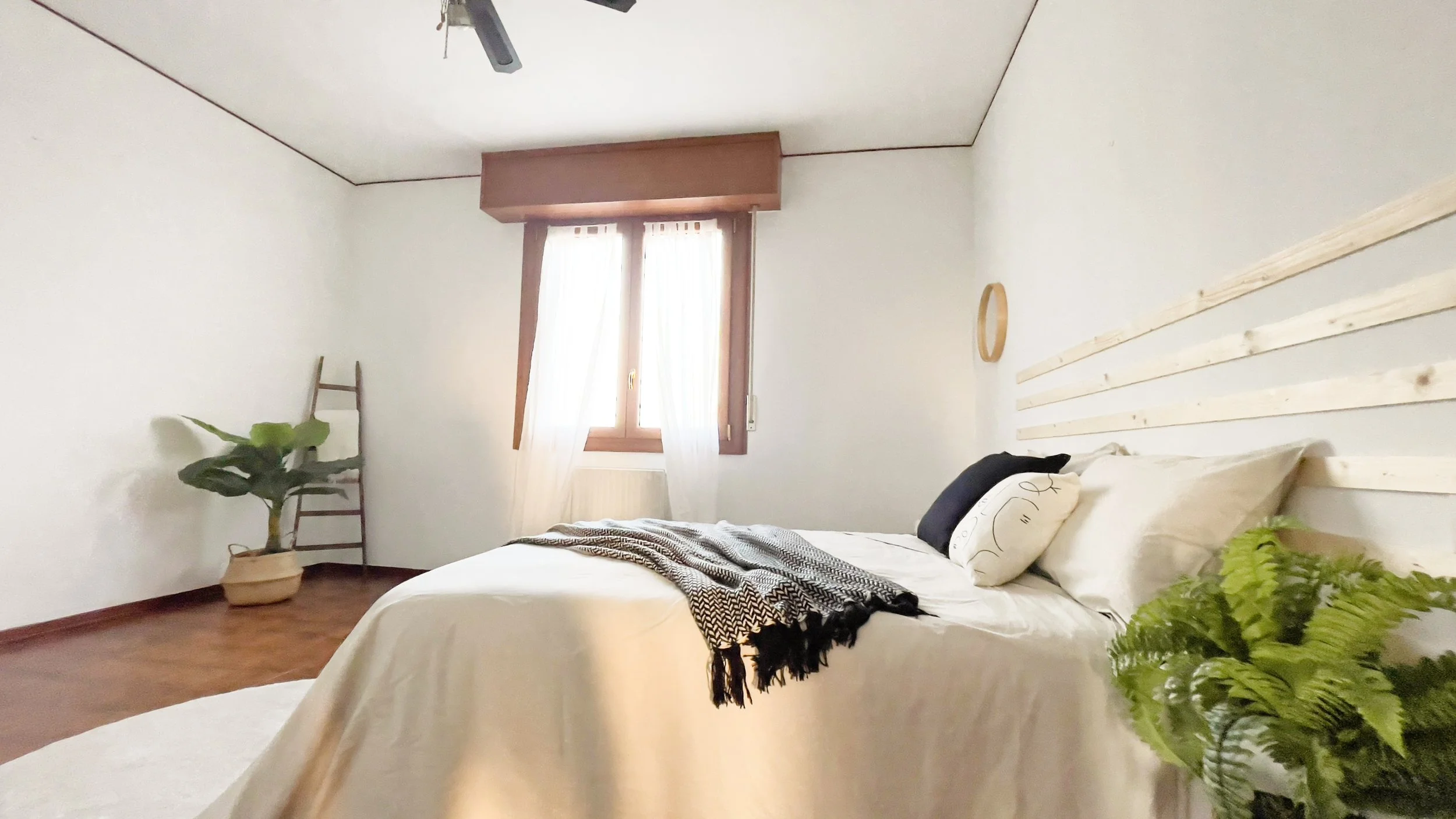 Camera da letto minimalista con letto matrimoniale, coperte e cuscini, pianta in vaso, scala decorativa in legno e una finestra con tende bianche.