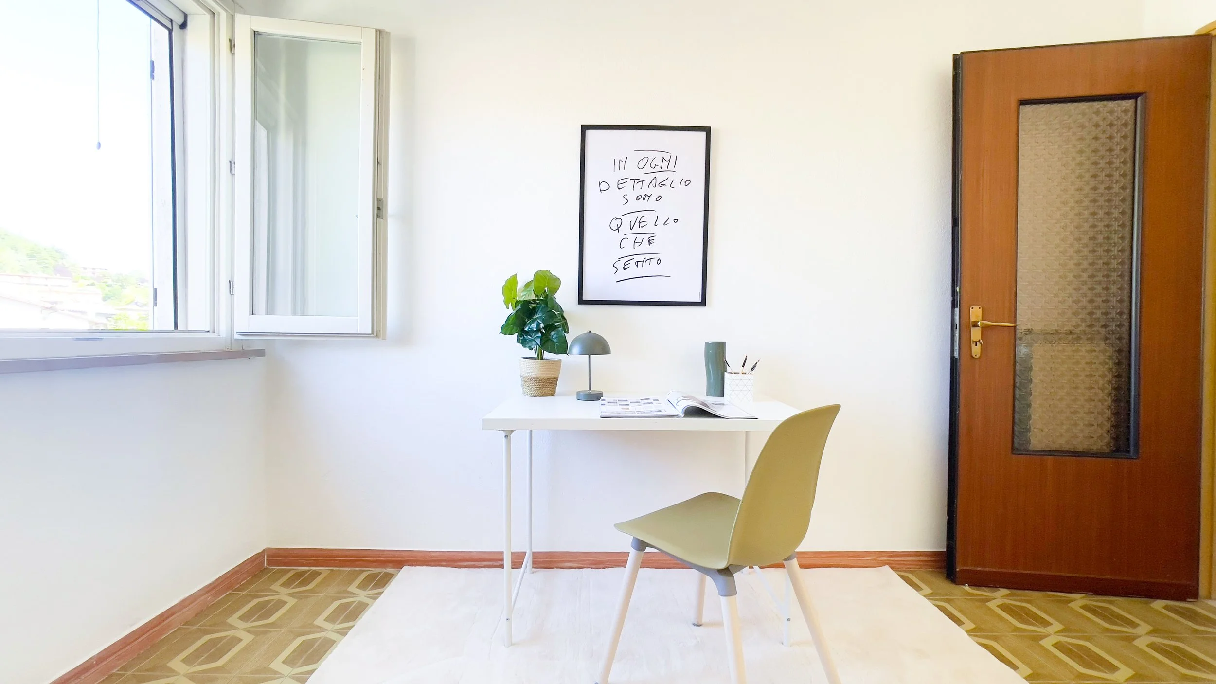 Interno di una stanza minimalista con una scrivania bianca, sedia verde, pianta, lampada, libri e finestra aperta.