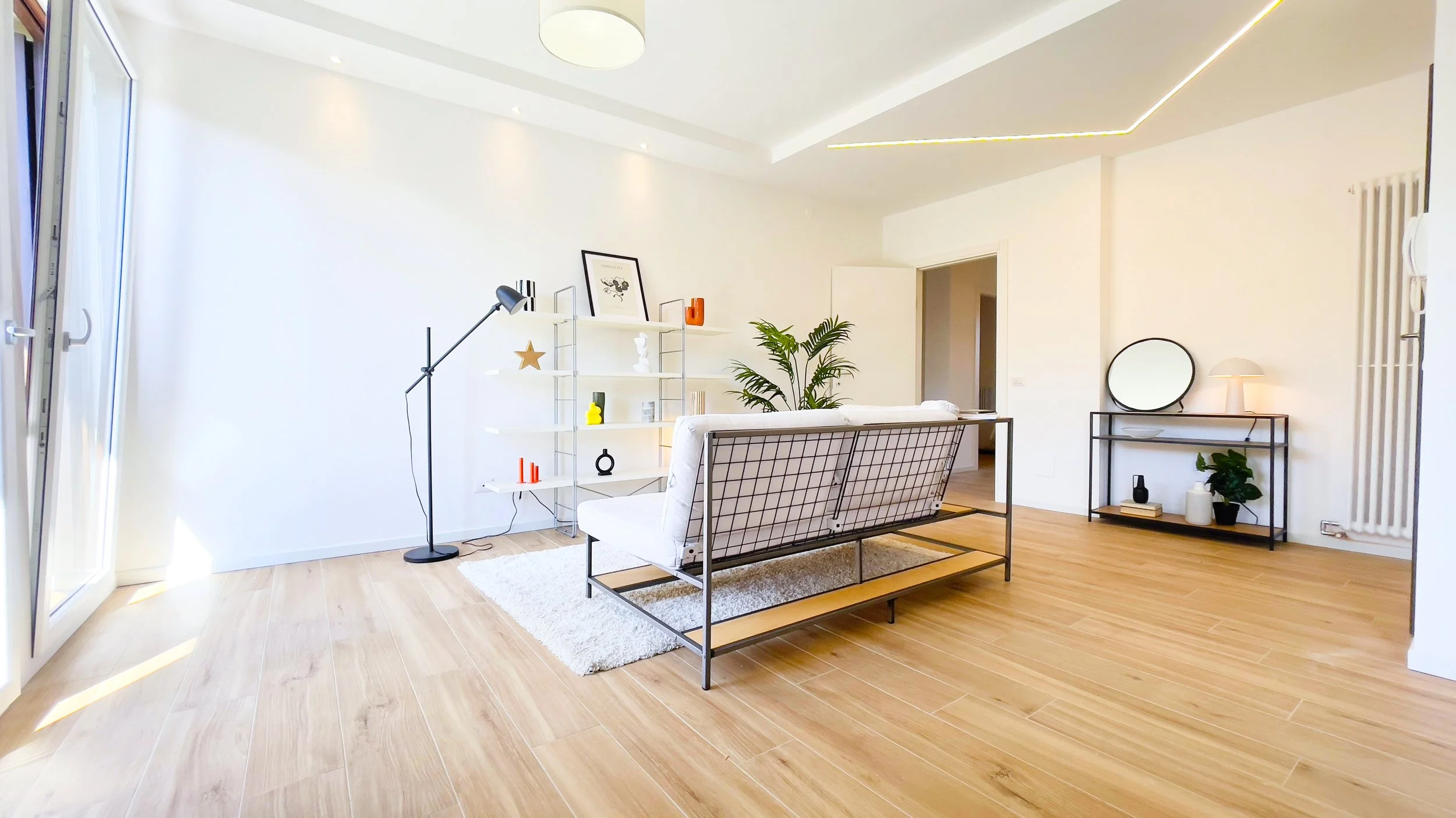 Soggiorno moderno con pareti bianche, pavimento in legno chiaro, divano con struttura in metallo, libreria con decorazioni, lampada da terra, mensola con piante e specchio.