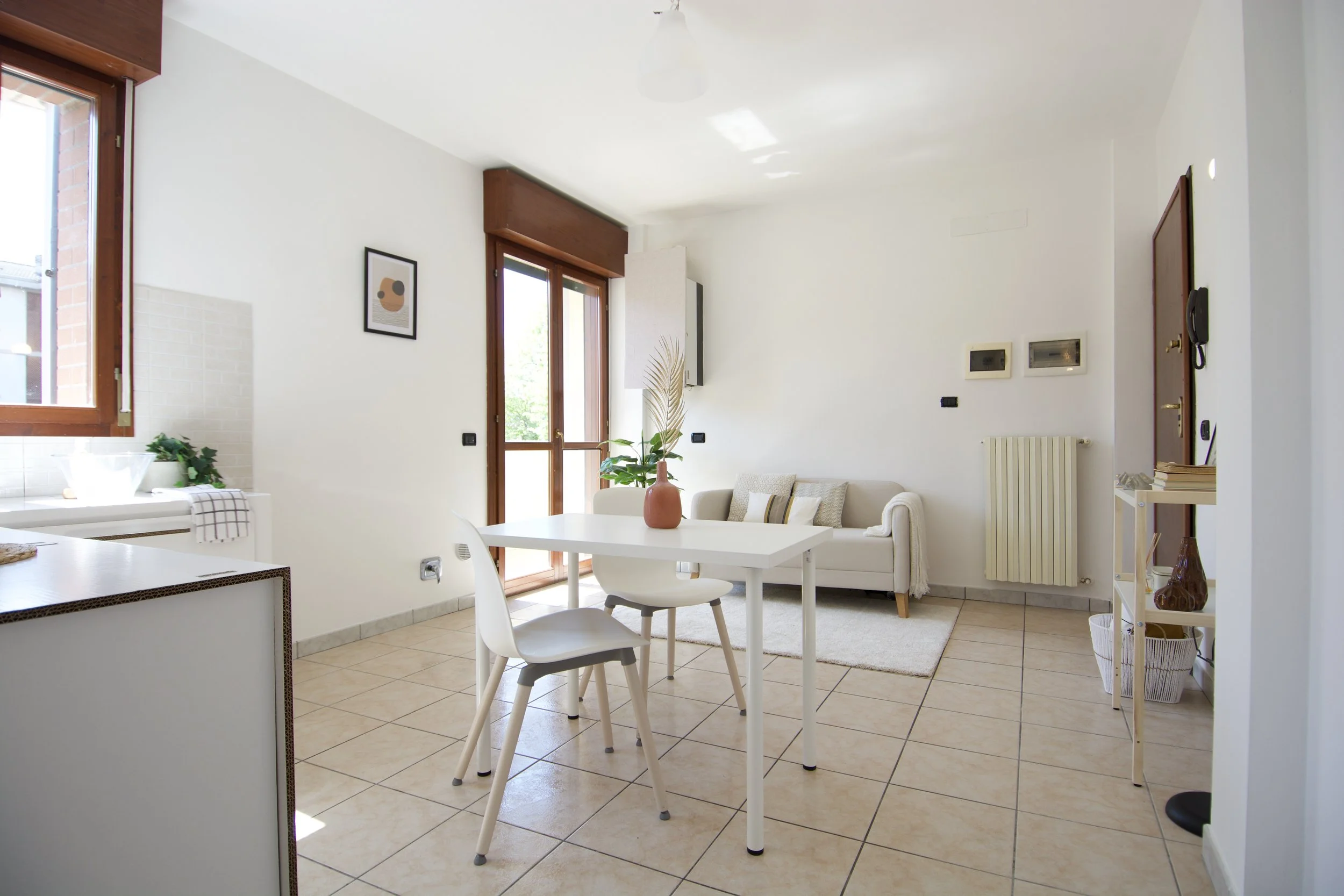 Interno di un appartamento moderno e luminoso con pavimento in piastrelle. La stanza ha una finestra con cornice in legno, un tavolo bianco con due sedie, una pianta in vaso sul tavolo, un divano bianco e alcune decorazioni alle pareti.