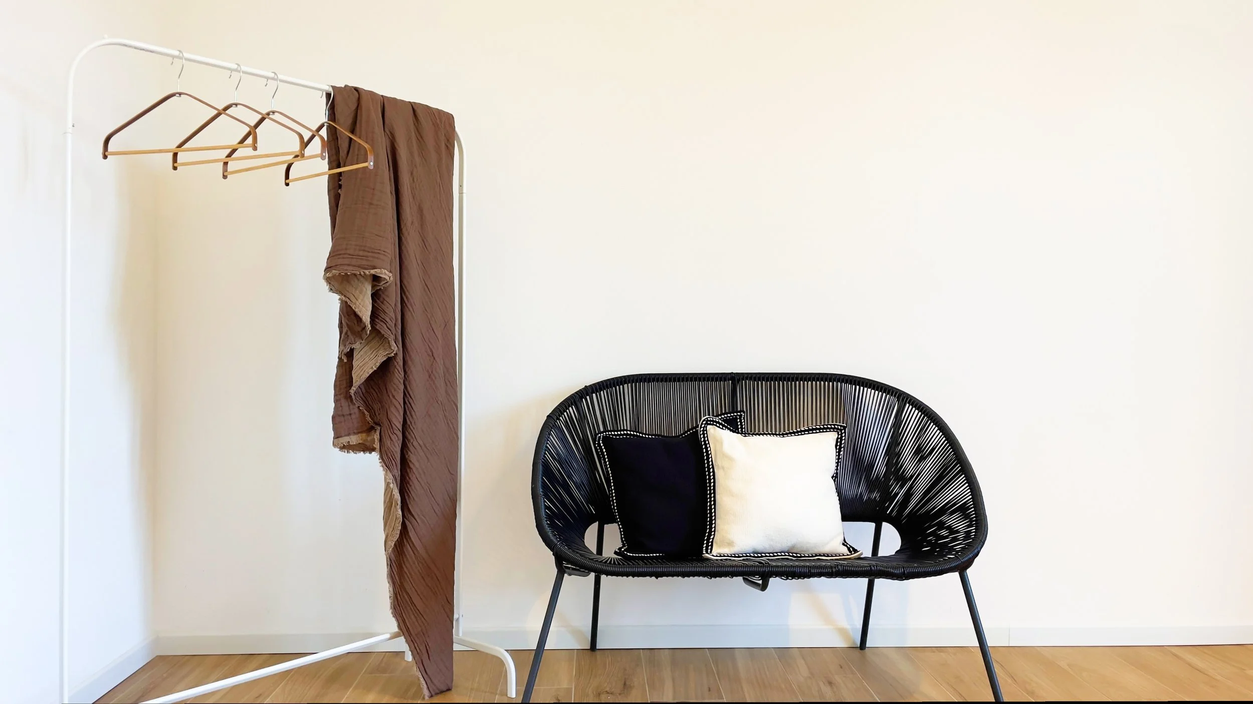 Sedia nera con cuscini bianchi e neri, accanto a un appendiabiti bianco con grucce e un panno marrone, su pavimento in legno.