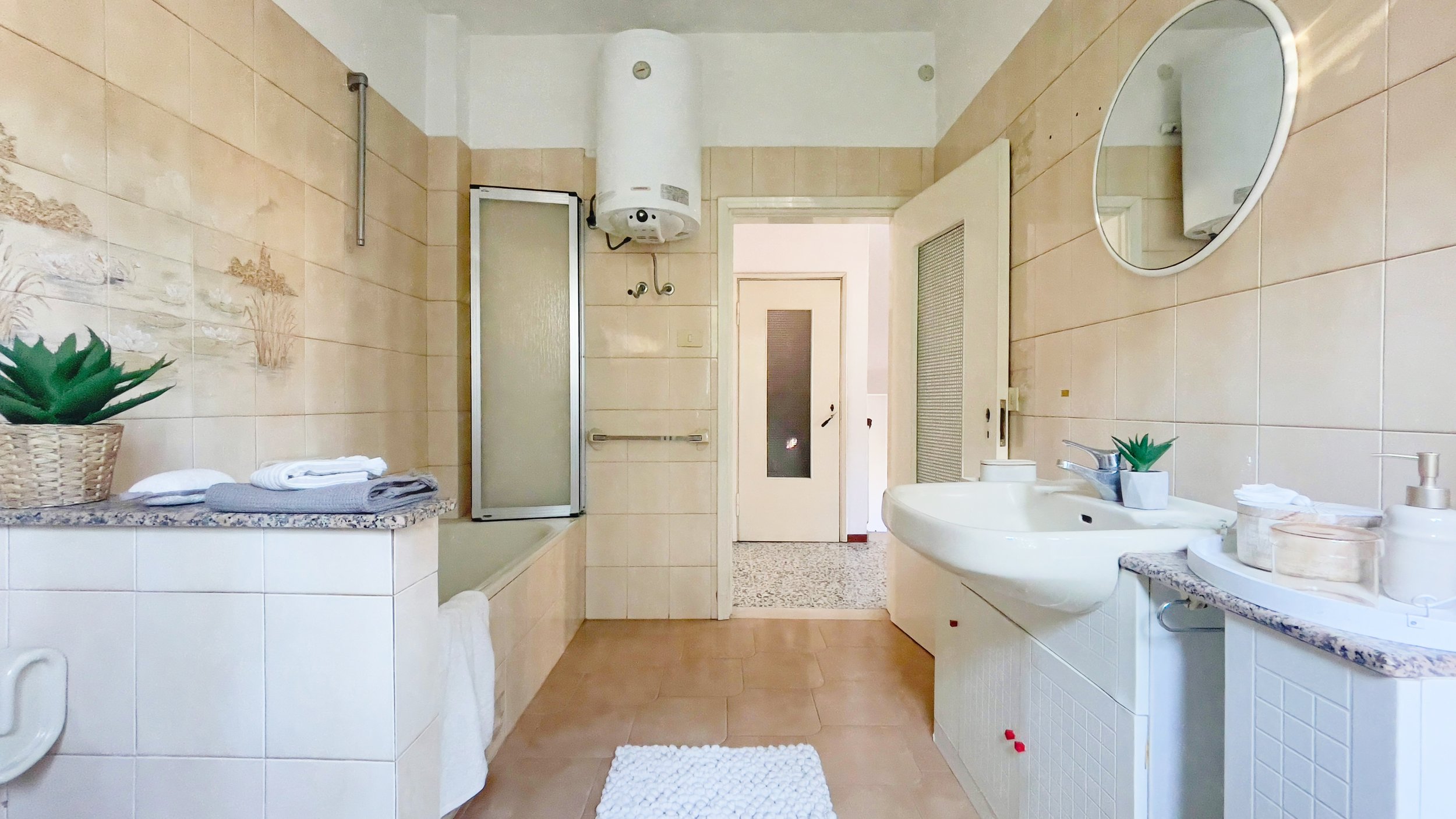 Bagno con vasca, lavabo, specchio rotondo, piastrelle beige, asciugamani piegati, piante in vaso e asciugamani sul bordo della vasca.