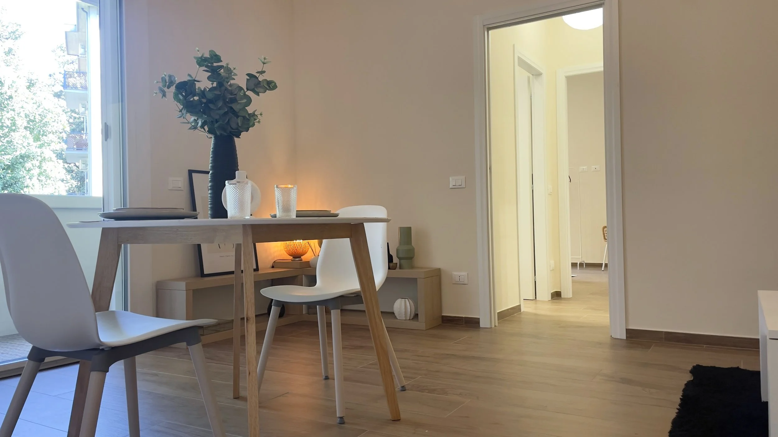Sala da pranzo moderna con tavolo in legno e sedie bianche, vaso con fiori su tavolo, pavimento in legno, porta aperta verso corridoio.