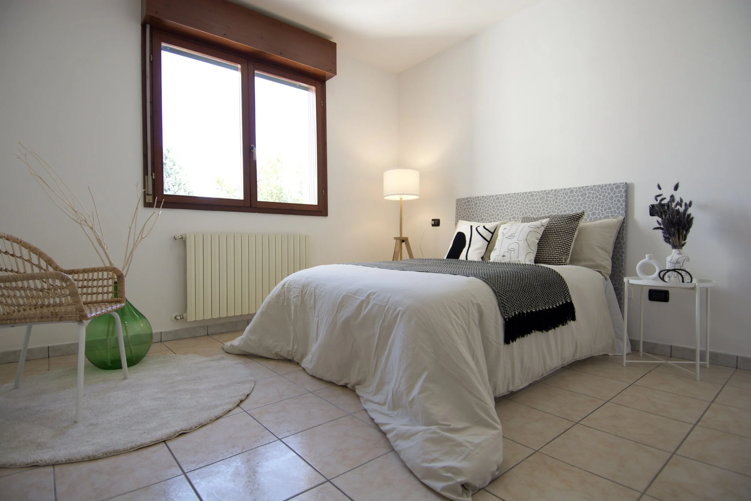 Camera da letto moderna con letto matrimoniale, lampada da terra, finestra con cornice in legno, sedia intrecciata, vaso verde e decorazioni sul comodino.