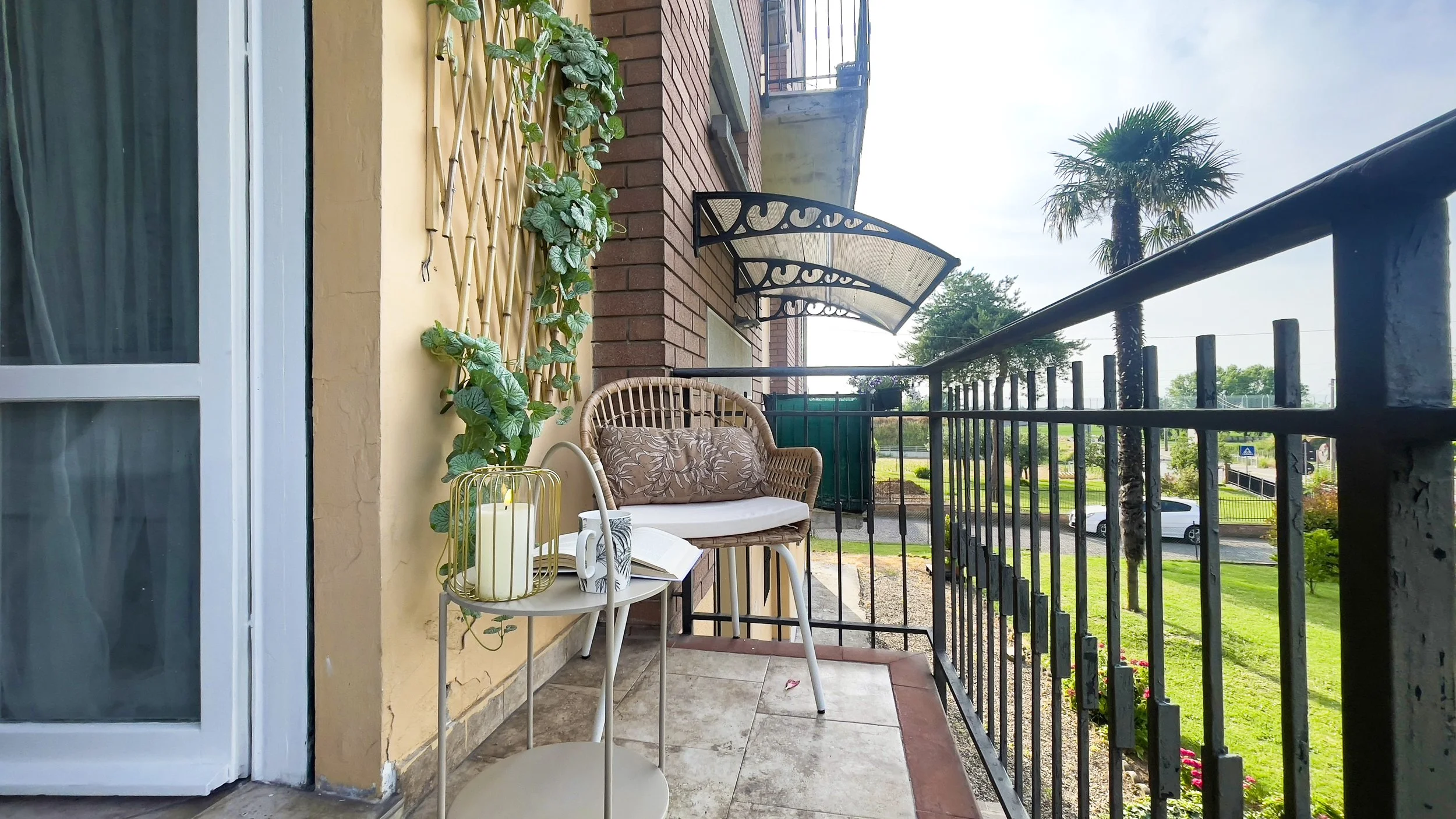 Balcone con sedia e tavolino, decorato con cuscini, piante rampicanti e candela, con vista sul giardino e la strada