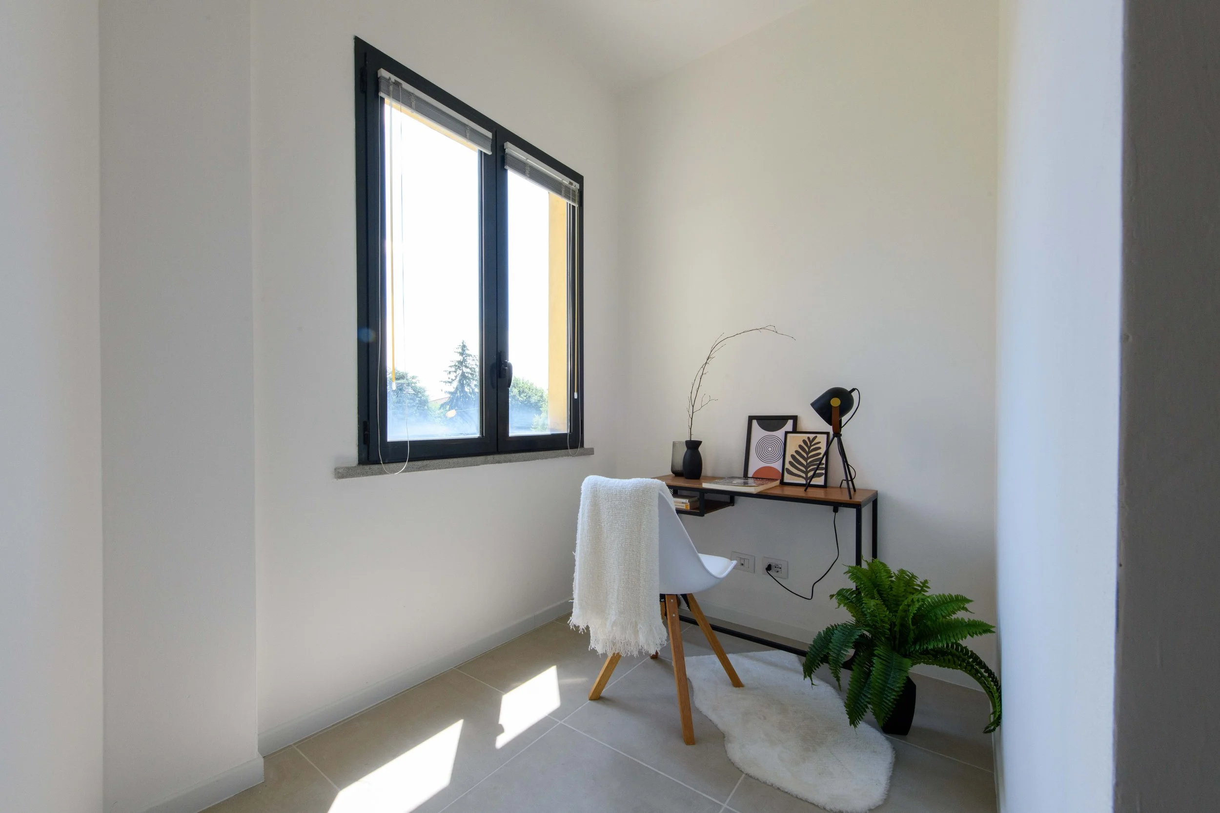 Piccola stanza luminosa con scrivania, sedia bianca, pianta verde, lampada, decorazioni e grande finestra.