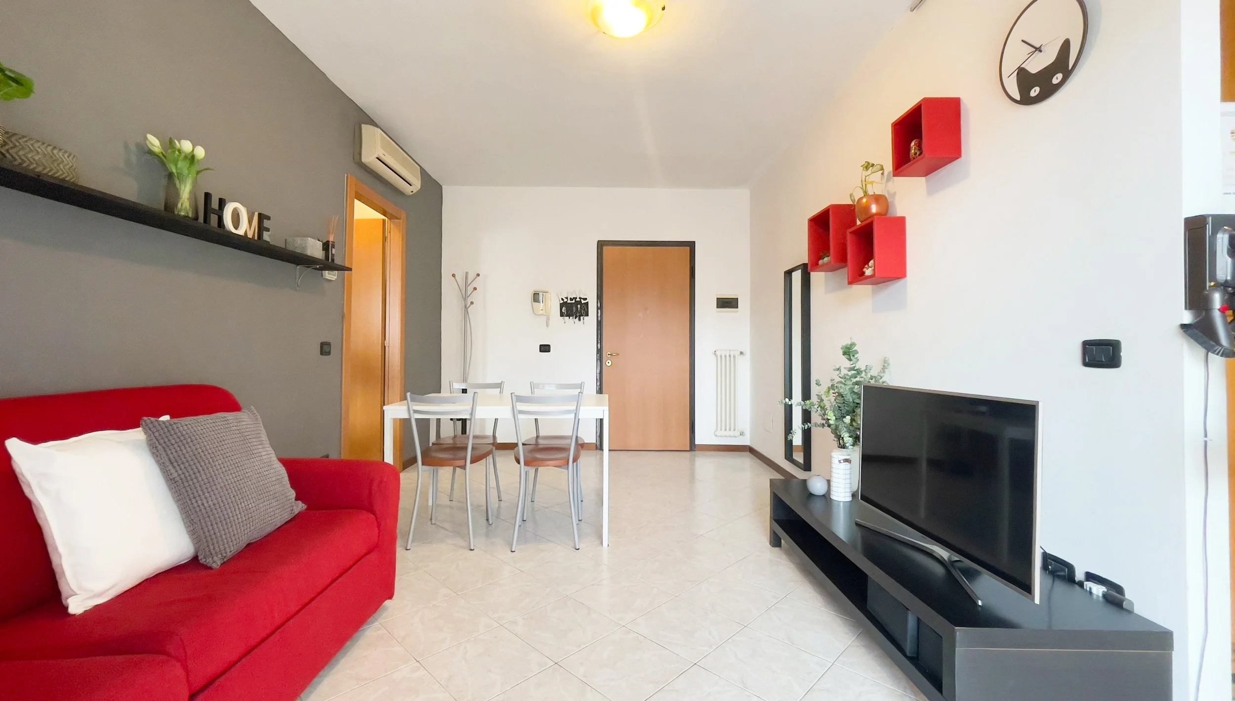 Soggiorno moderno con divano rosso, tavolo da pranzo bianco con sedie, parete grigia con mensola e decorazioni, TV su mobile nero, orologio a muro, e scaffali rossi.