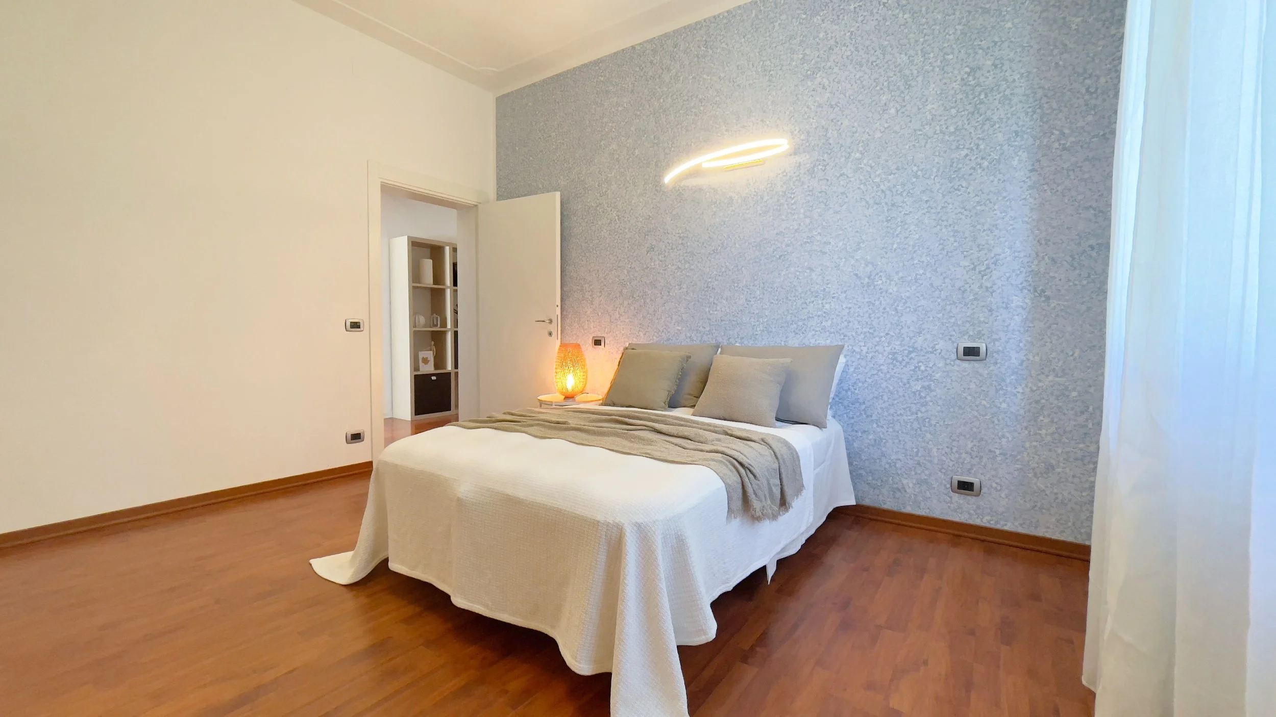 Camera da letto minimalista con letto matrimoniale, cuscini grigi, lampada accesa su un comodino, pareti bianche e pavimento in legno.