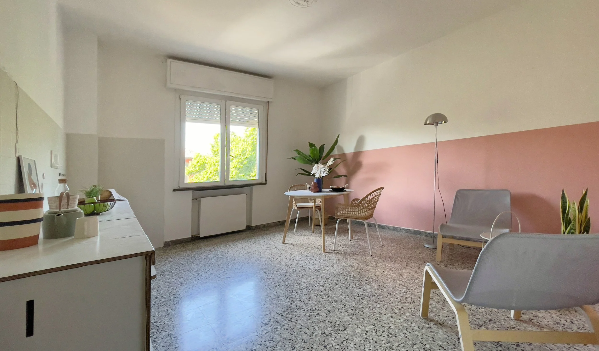 Salotto moderno con pareti bianche e rosa, una finestra luminosa, sedie in vimini intorno a un tavolo rotondo, una poltrona grigia, lampada ad arco e piante verdi sul pavimento di marmo.