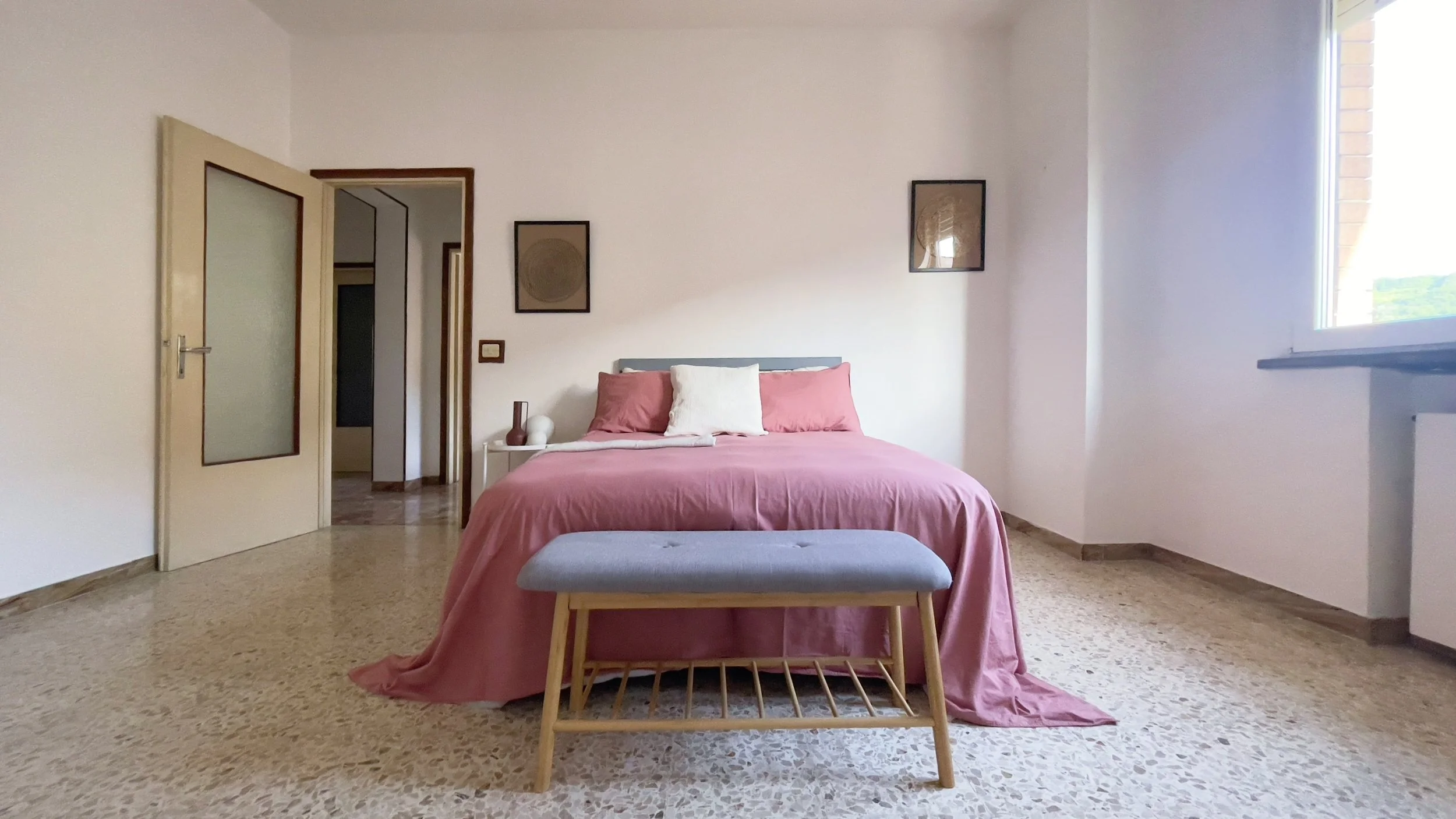 Camera da letto con letto matrimoniale rosa, panca ai piedi del letto, porta aperta a sinistra e finestra a destra.