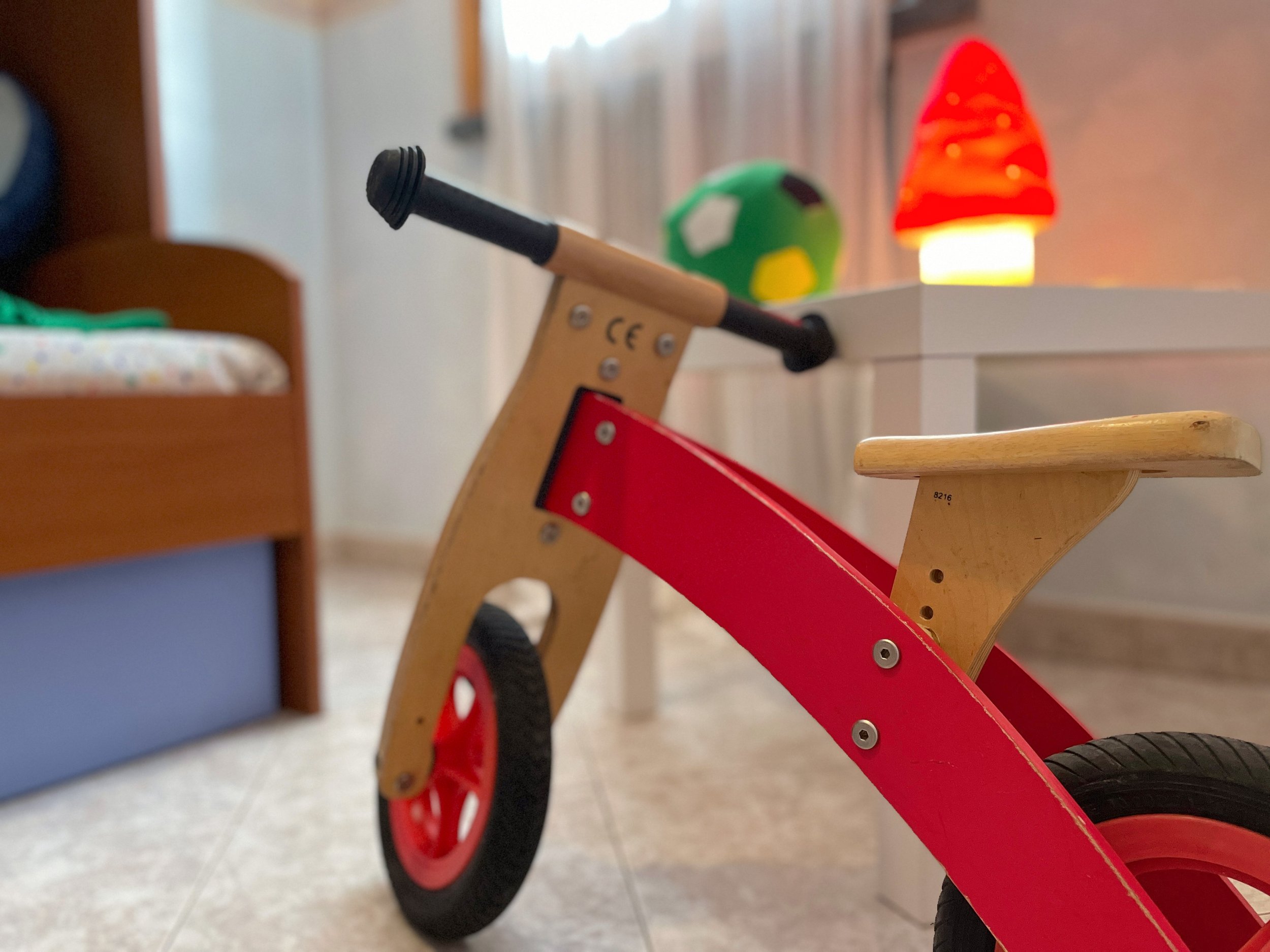 Bicicletta senza pedali in legno e rosso in una stanza da bambini con letto, tavolo e un giocattolo a forma di lampada fungo sullo sfondo.