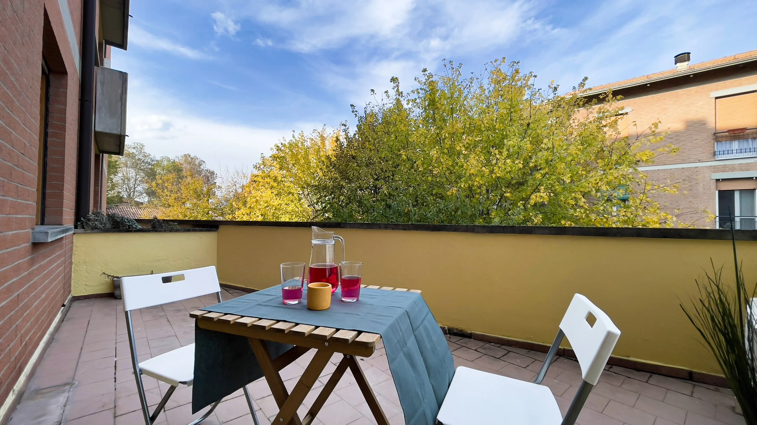 Terrazza con tavolo di legno, due sedie bianche, caraffa e bicchieri con bevanda rossa, sfondo di alberi e cielo soleggiato.