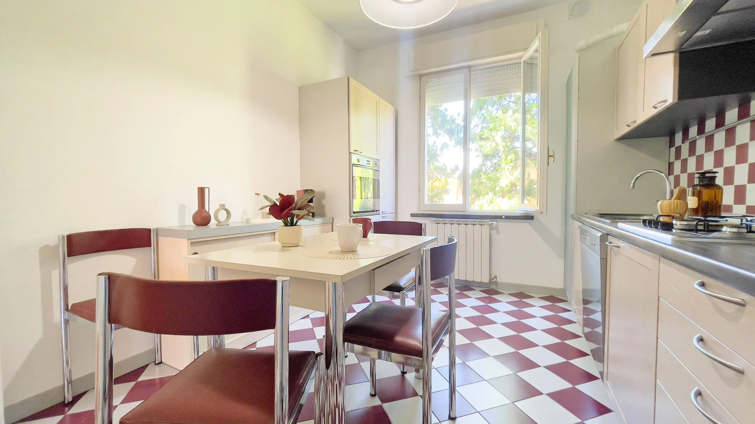 Cucina moderna con tavolo bianco, sedie marroni, pavimento a scacchi rosso e bianco, finestra aperta, e mobili chiari.