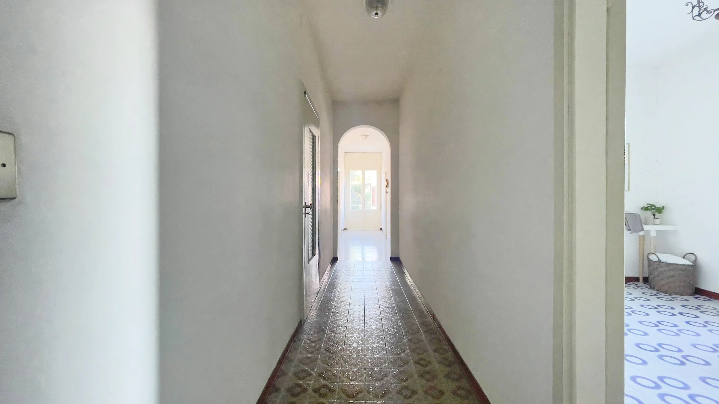 Un corridoio lungo e stretto con pavimento a piastrelle e un arco che conduce a una stanza luminosa con una finestra in fondo. A destra, una porta aperta mostra una stanza laterale con un pavimento a motivi geometrici e un cestino grigio in un angolo