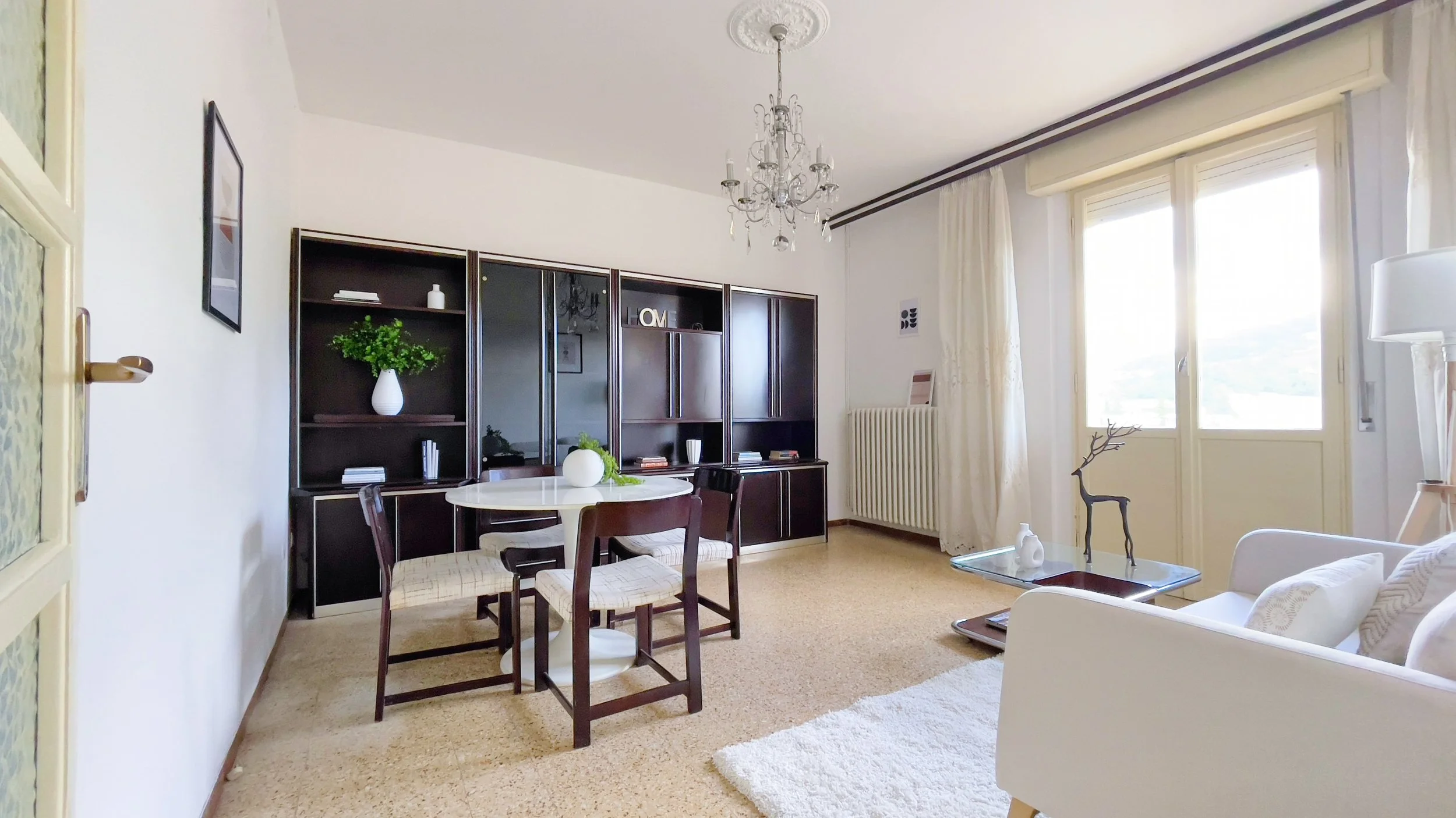 Soggiorno elegante con tavolo da pranzo rotondo e sedie, libreria nera, divano bianco, tappeto chiaro, lampadario di cristallo e porta finestra.