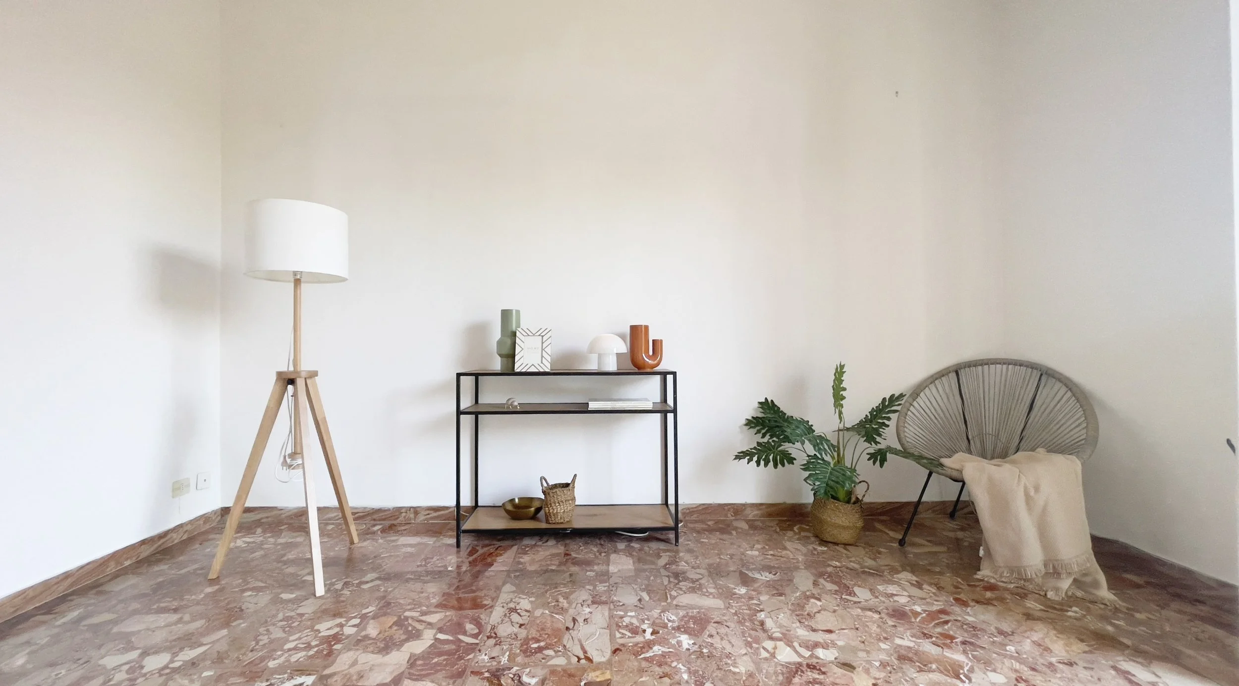 Interno di una stanza minimalista con pavimento in marmo. A sinistra, una lampada da terra in legno. Al centro, una mensola con oggetti decorativi e un piccolo vaso di piante. A destra, una sedia con una coperta beige appoggiata sopra.