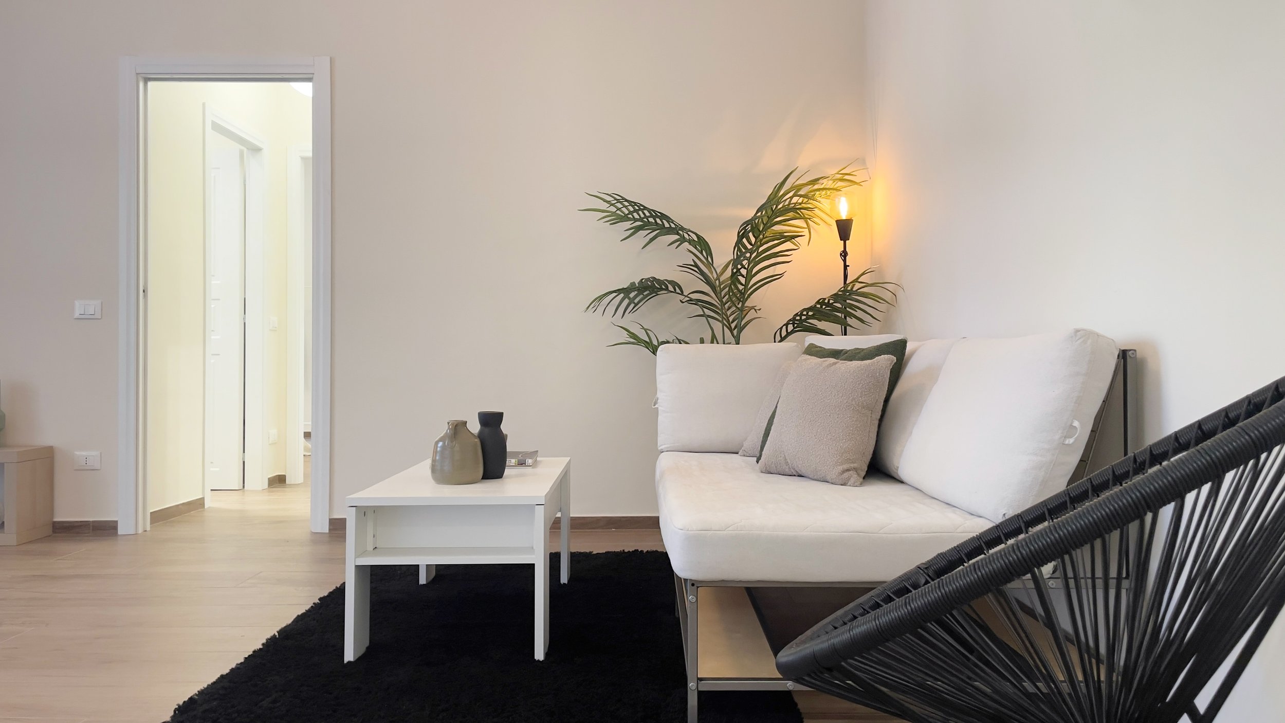 Soggiorno moderno con divano bianco, tavolino bianco, cuscini, lampada, pianta verde e tappeto nero.