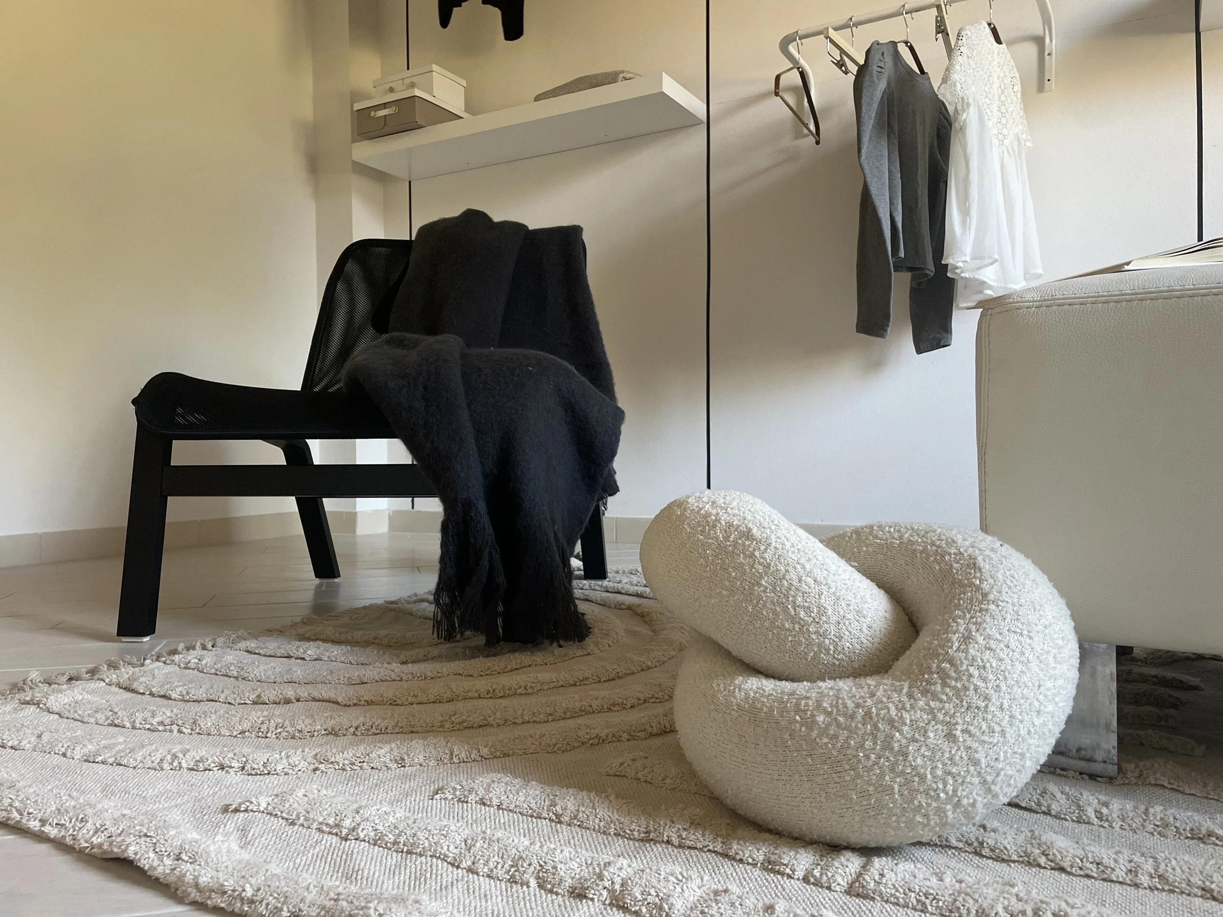 Interno di una stanza con sedia nera, coperta scura sopra, tappeto chiaro, cuscino intrecciato beige sul pavimento, scaffale bianco con scatole, abiti appesi su una barra.