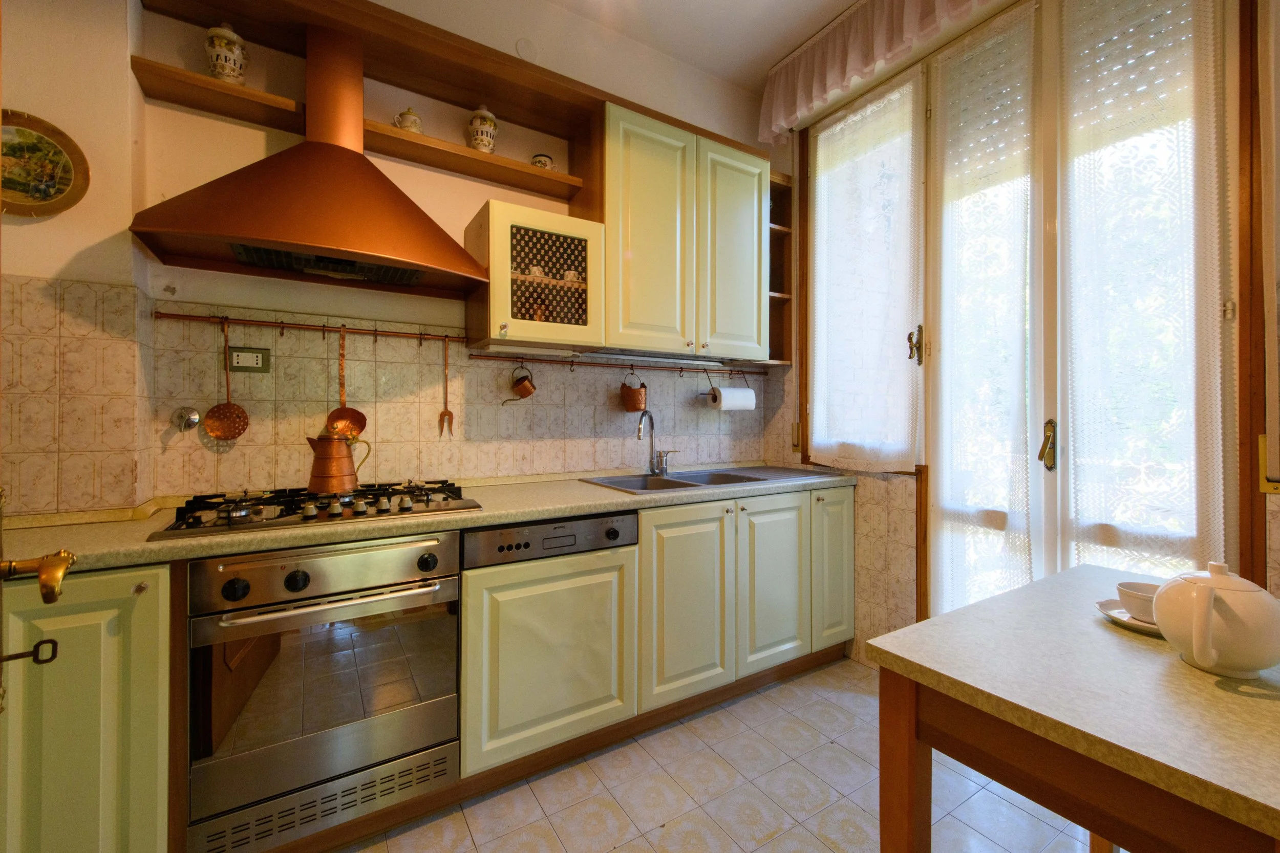 Cucina tradizionale con mobili color crema e cappa in rame, piano cottura e forno, lavello accanto a finestra con tende bianche, tavolo con teiera e tazze.