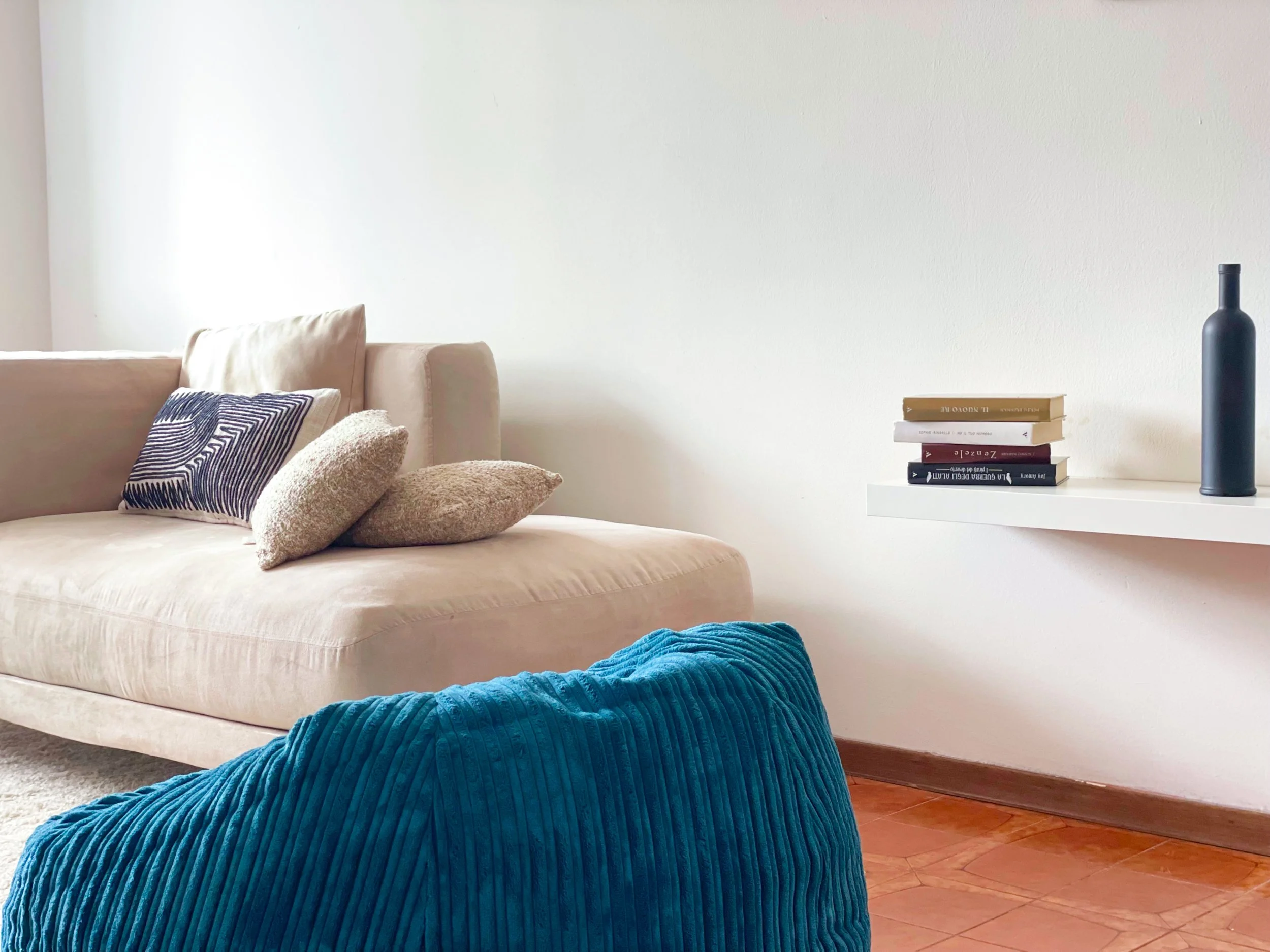 Soggiorno moderno con divano beige, cuscini decorativi e pouf blu; mensola con libri e bottiglia decorativa nera.