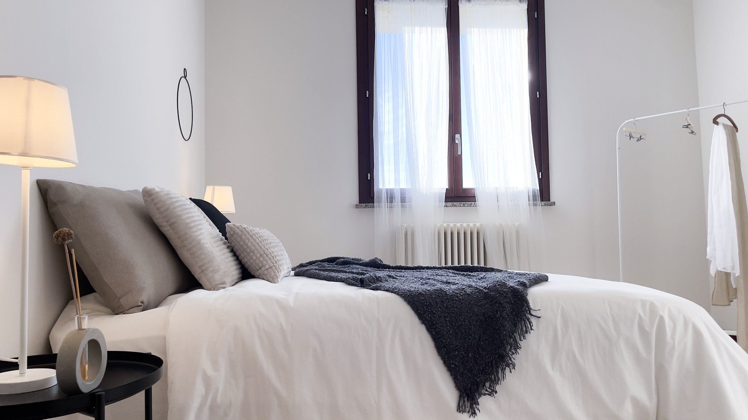 Camera da letto moderna con letto matrimoniale, cuscini grigi, coperta scura, lampada, finestra con tende bianche e porta abiti.