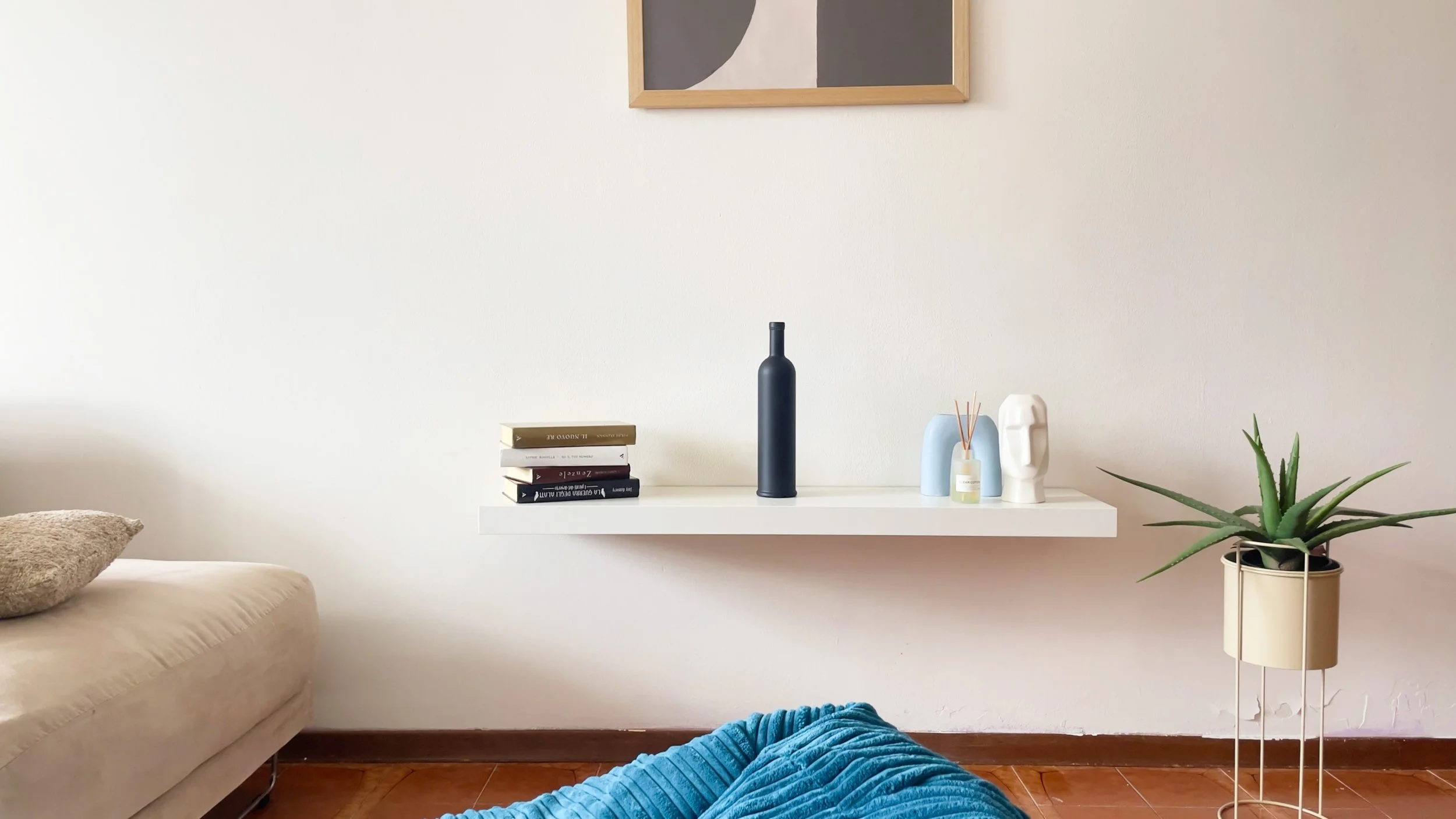 Salotto moderno con divano beige, mensola bianca con libri e decorazioni, un vaso con bastoncini profumati, pianta verde in vaso e quadro minimalista appeso alla parete.