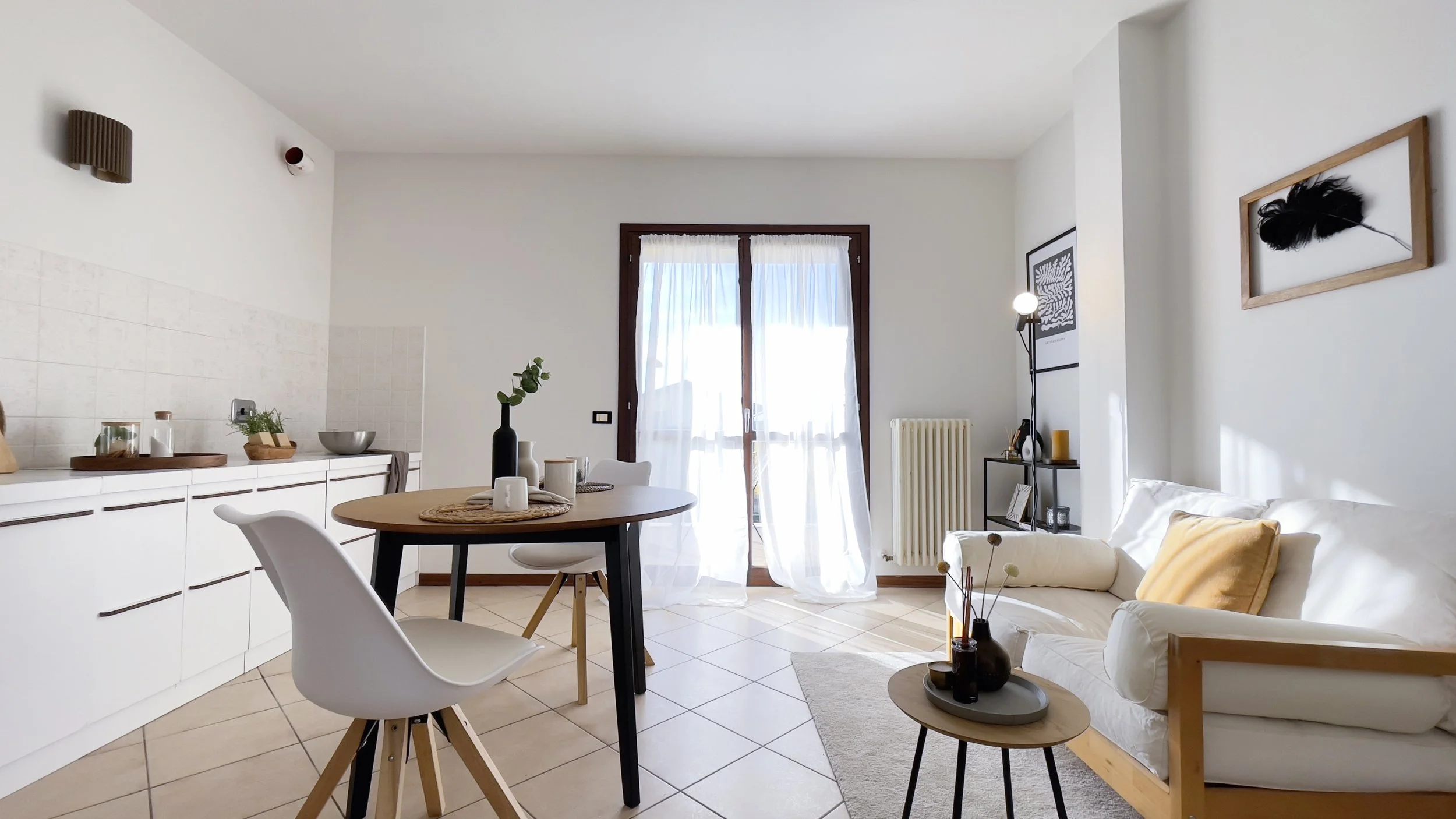 Salotto luminoso con arredamento minimalista, tavolo e sedie bianche, divano bianco con cuscini beige, piante decorative su un tavolino e grande finestra con tende.