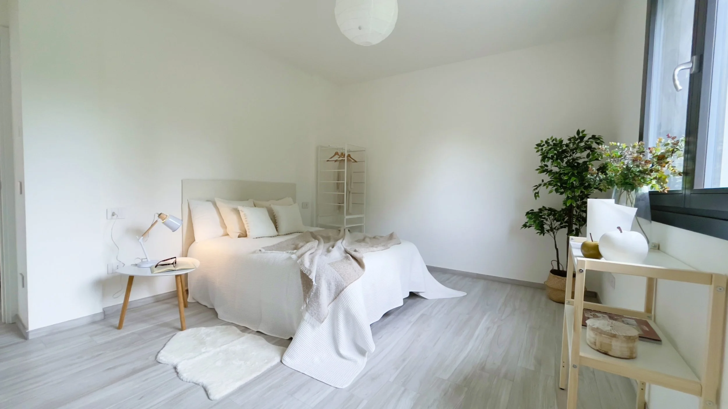 Camera da letto moderna con letto matrimoniale bianco, coperta beige, comodino con lampada e libro, tappeto soffice, appendiabiti, pianta d'appartamento e finestra con luce naturale.