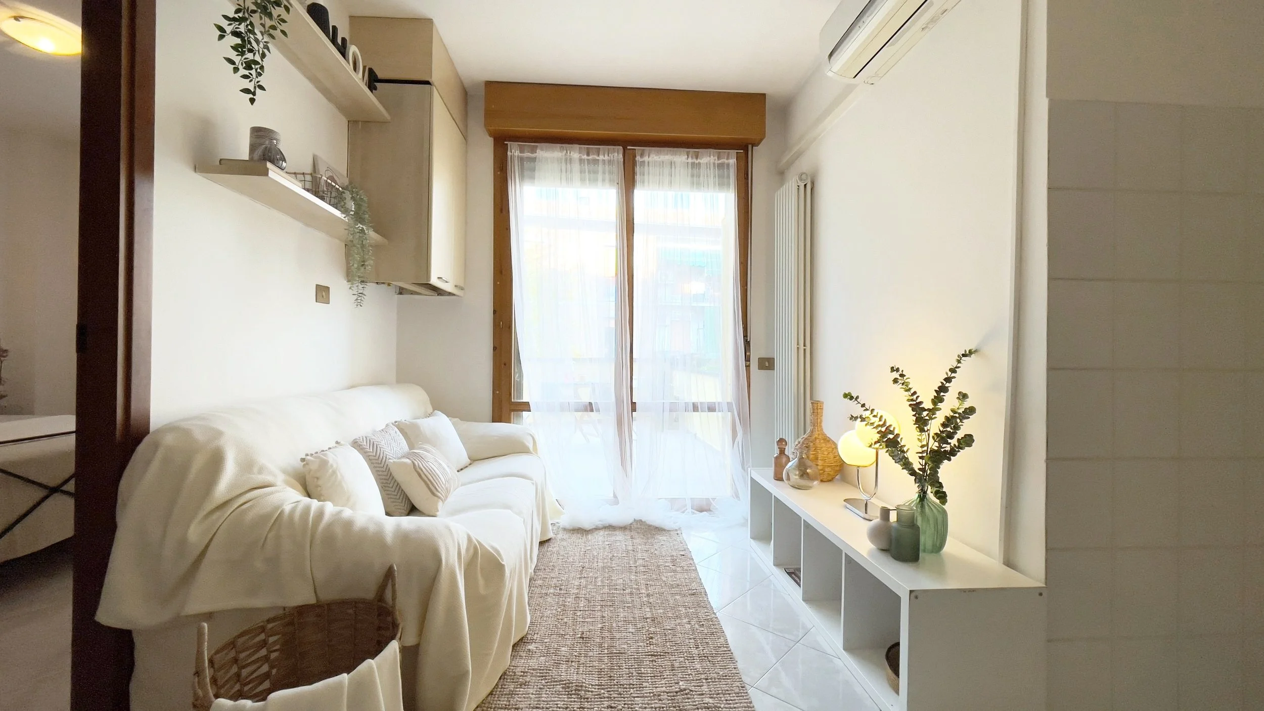 Soggiorno luminoso con divano bianco, tappeto beige e decorazioni naturali. Tavolino con piante, lampada e bottiglie di vetro su scaffale bianco. Tende trasparenti su ampia finestra. Aria condizionata sul muro.