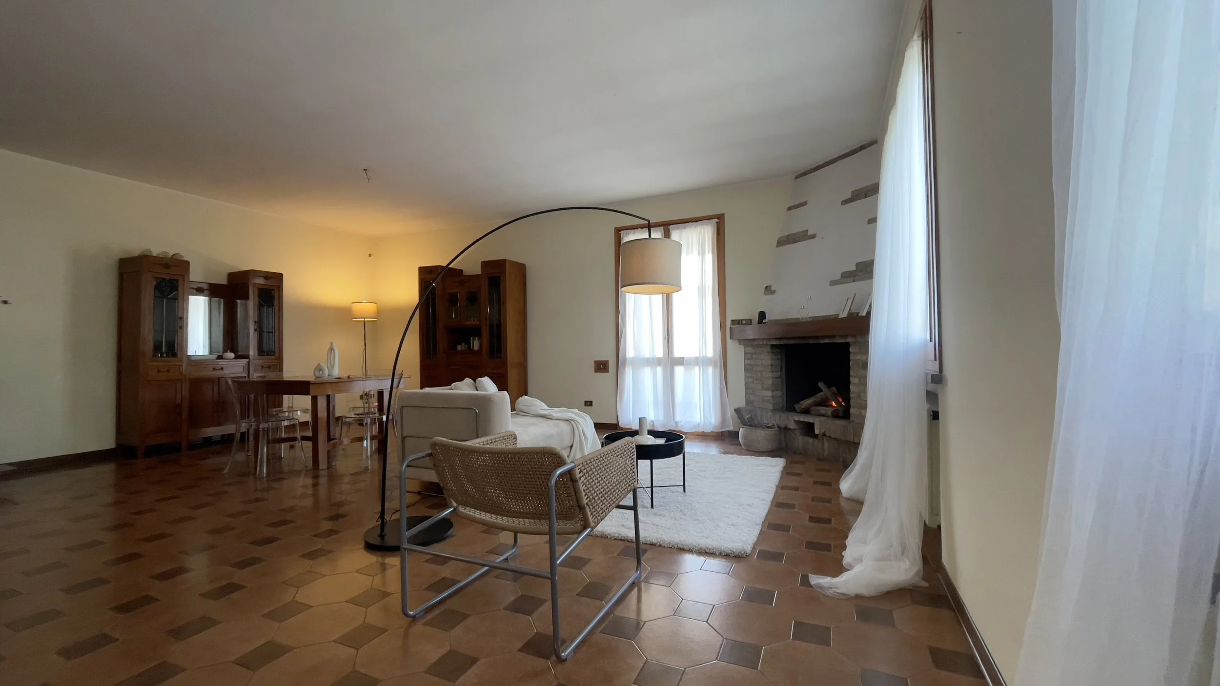 Soggiorno con arredamento rustico, caminetto acceso, poltrona con seduta intrecciata, tavolo in legno, e grande lampada ad arco. Finestre con tendaggi bianchi chiari.