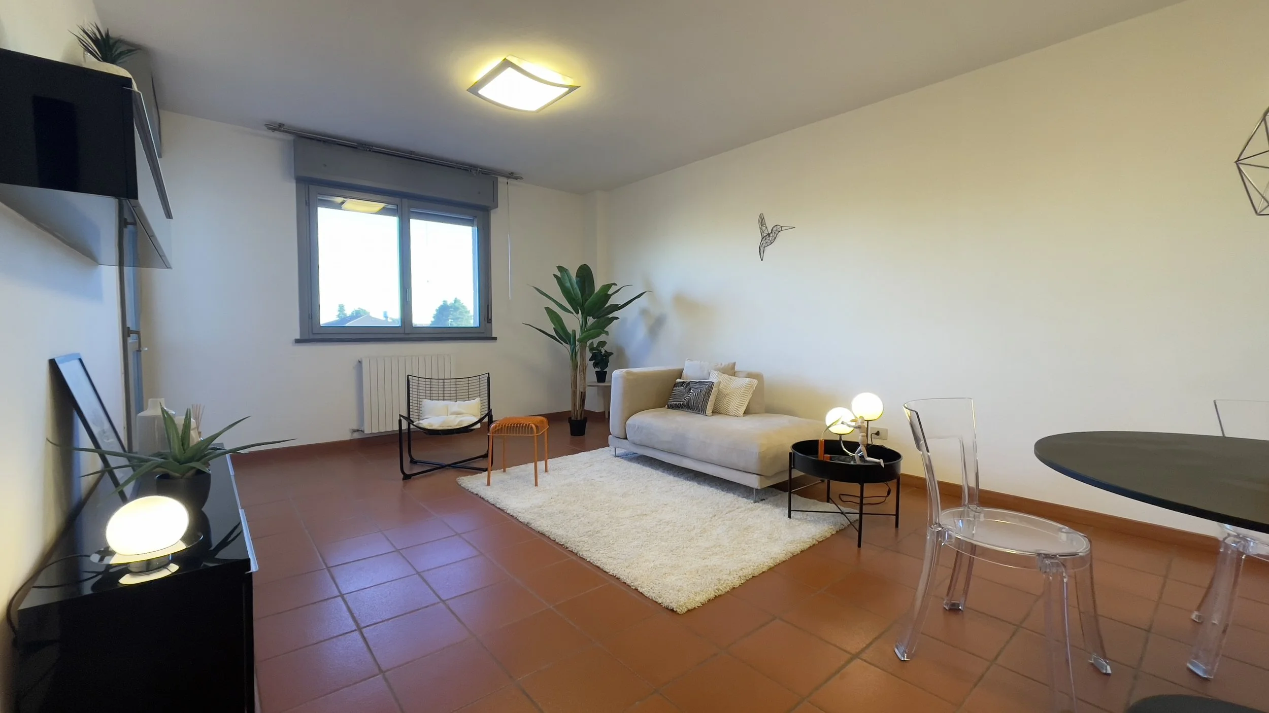 Soggiorno moderno con divano beige, tappeto bianco, poltrona nera, tavolino, piante decorative e pavimento in terracotta.