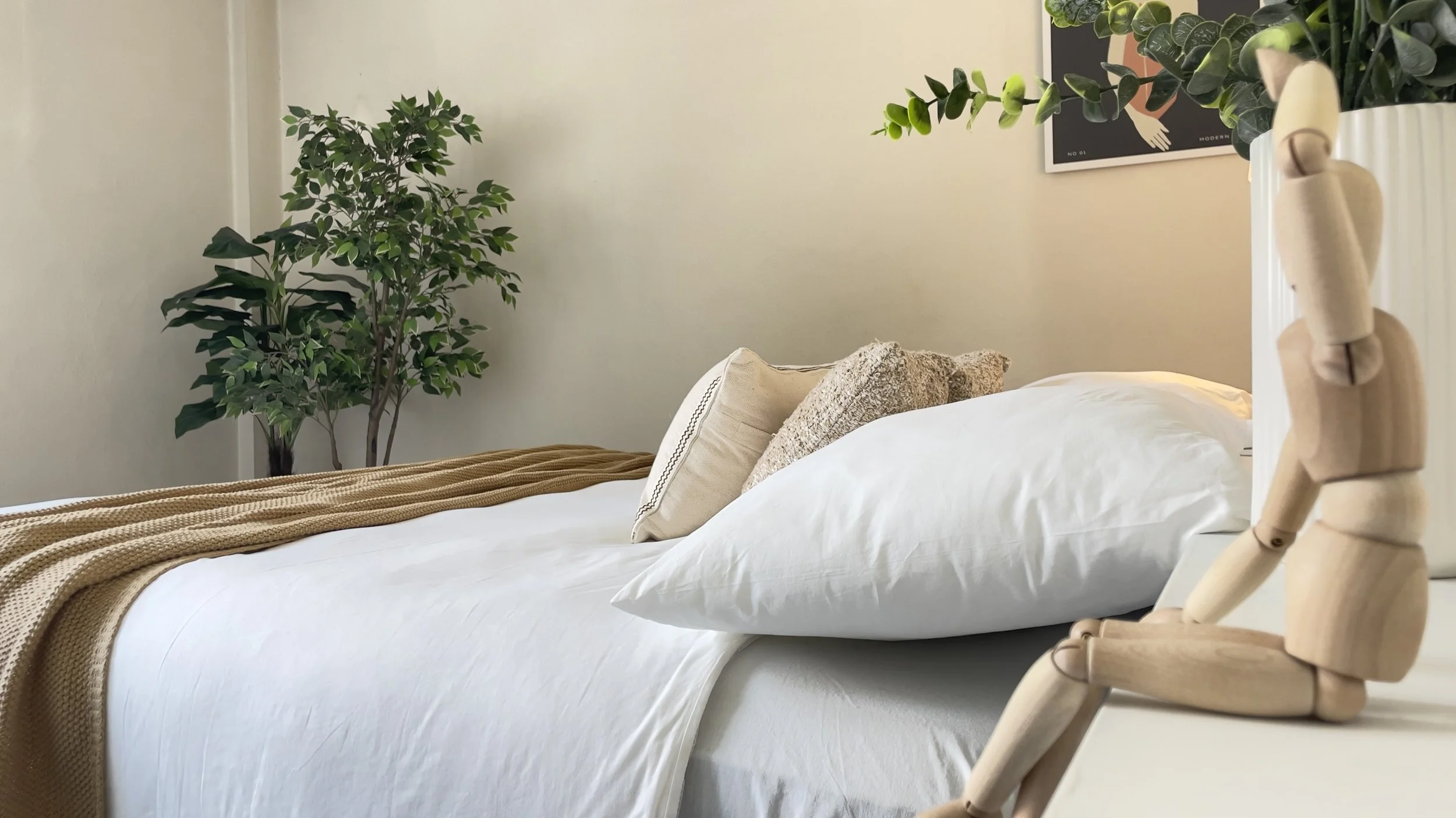 Camera da letto moderna con letto bianco e cuscini, piante decorative e manichino di legno su una mensola.