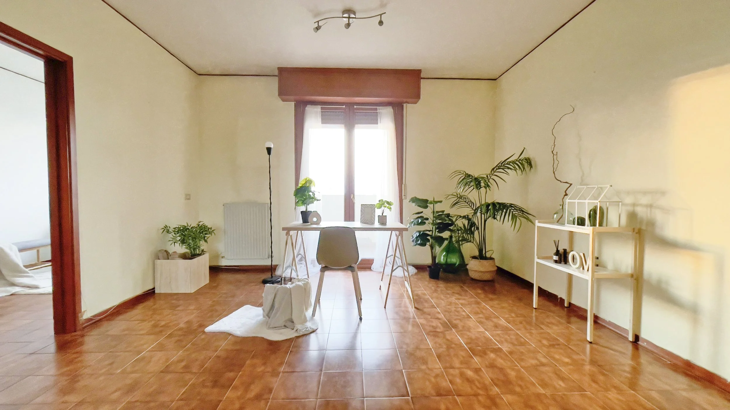 Interno di una stanza luminosa con pavimento in legno, tavolo e sedia al centro, piante verdi, mensola bianca con decorazioni e ampia finestra.