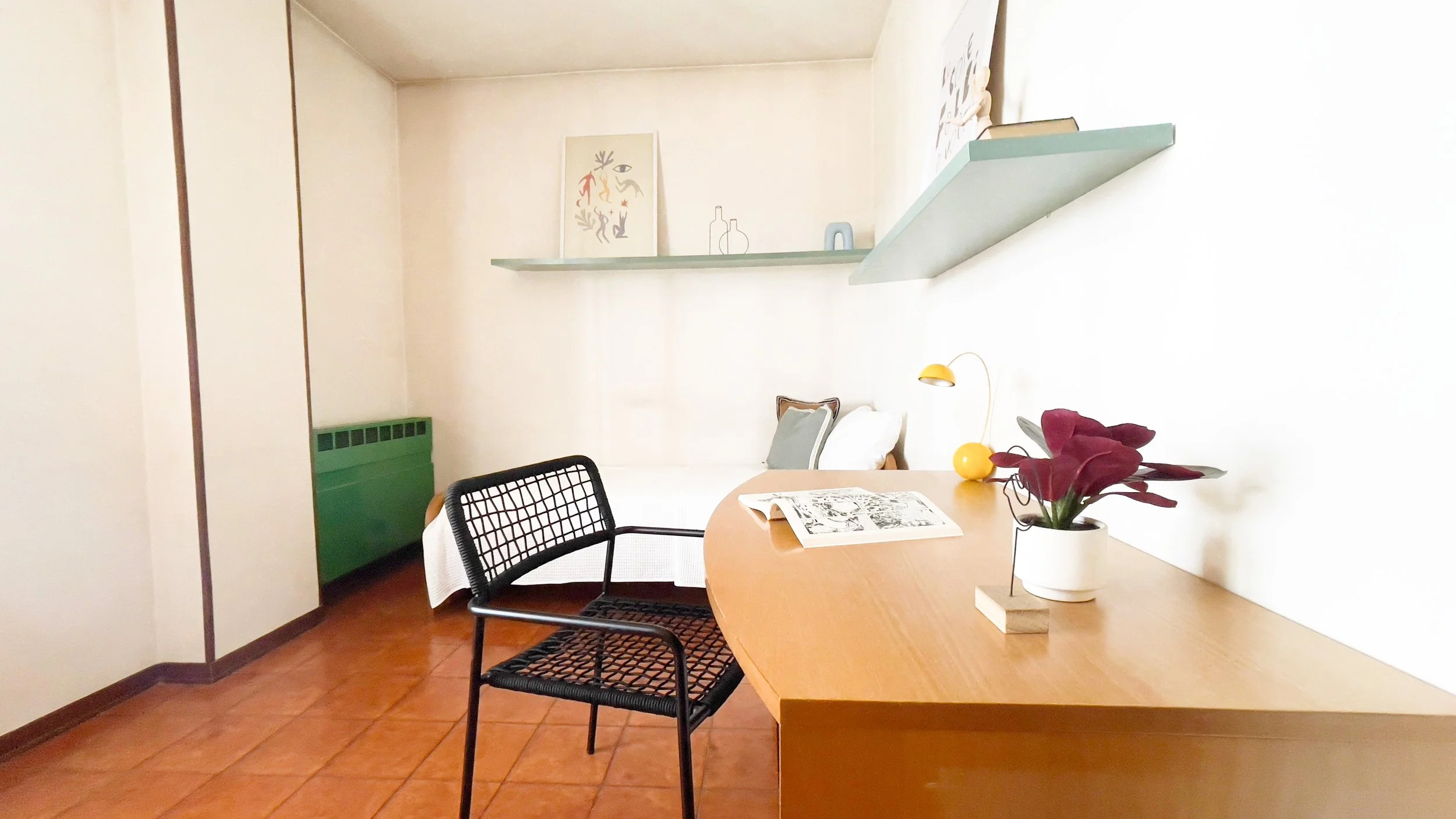 Studio minimalista con tavolo in legno, sedia nera, pianta decorativa e lampada gialla. Un letto si intravede sullo sfondo, con cuscini e una mensola con decorazioni appesa sopra. La stanza ha pavimento in piastrelle arancioni e pareti chiare.