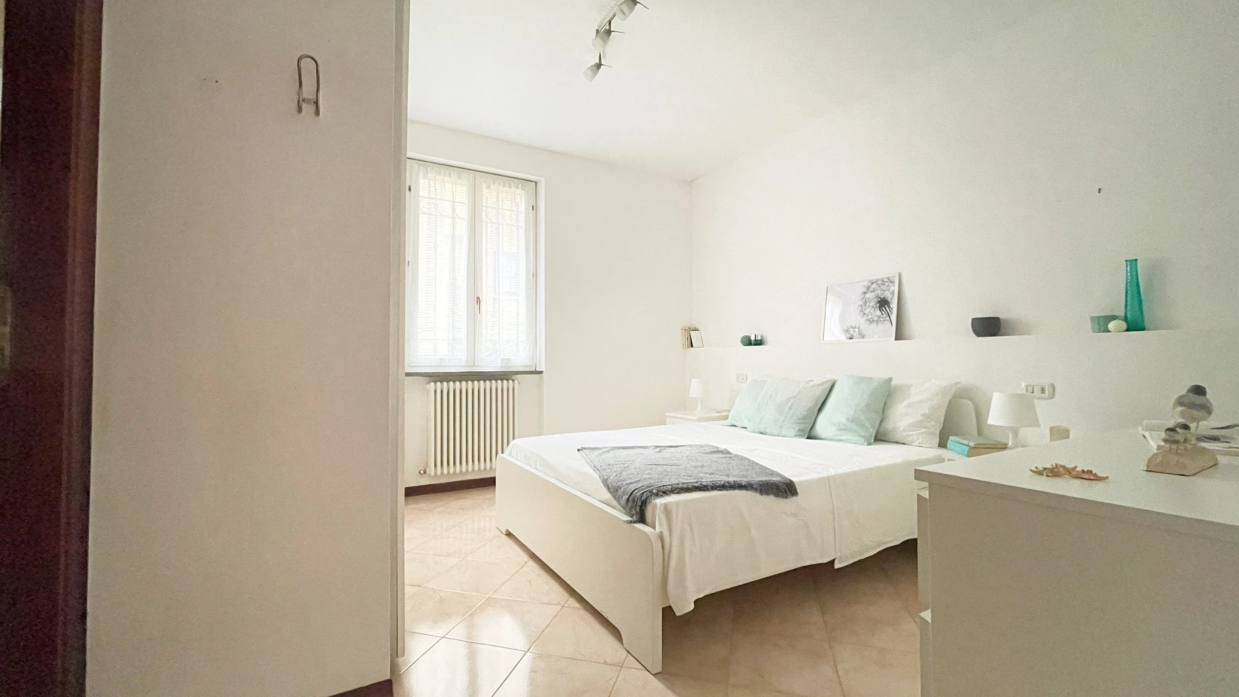 Camera da letto moderna e minimalista con letto matrimoniale, cuscini color pastello, coperta grigia e decorazioni semplici su mensole.