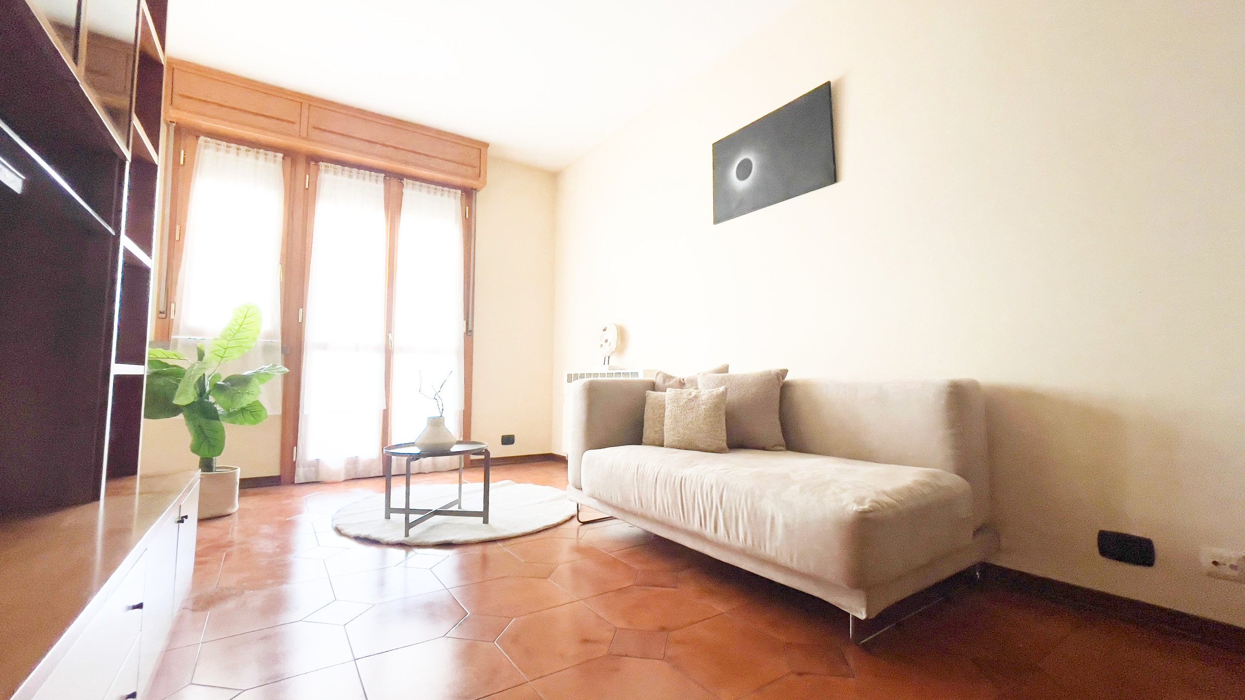 Soggiorno luminoso con divano beige, tavolino, pianta, grande finestra, pavimento in piastrelle marroni e quadro nero sul muro.
