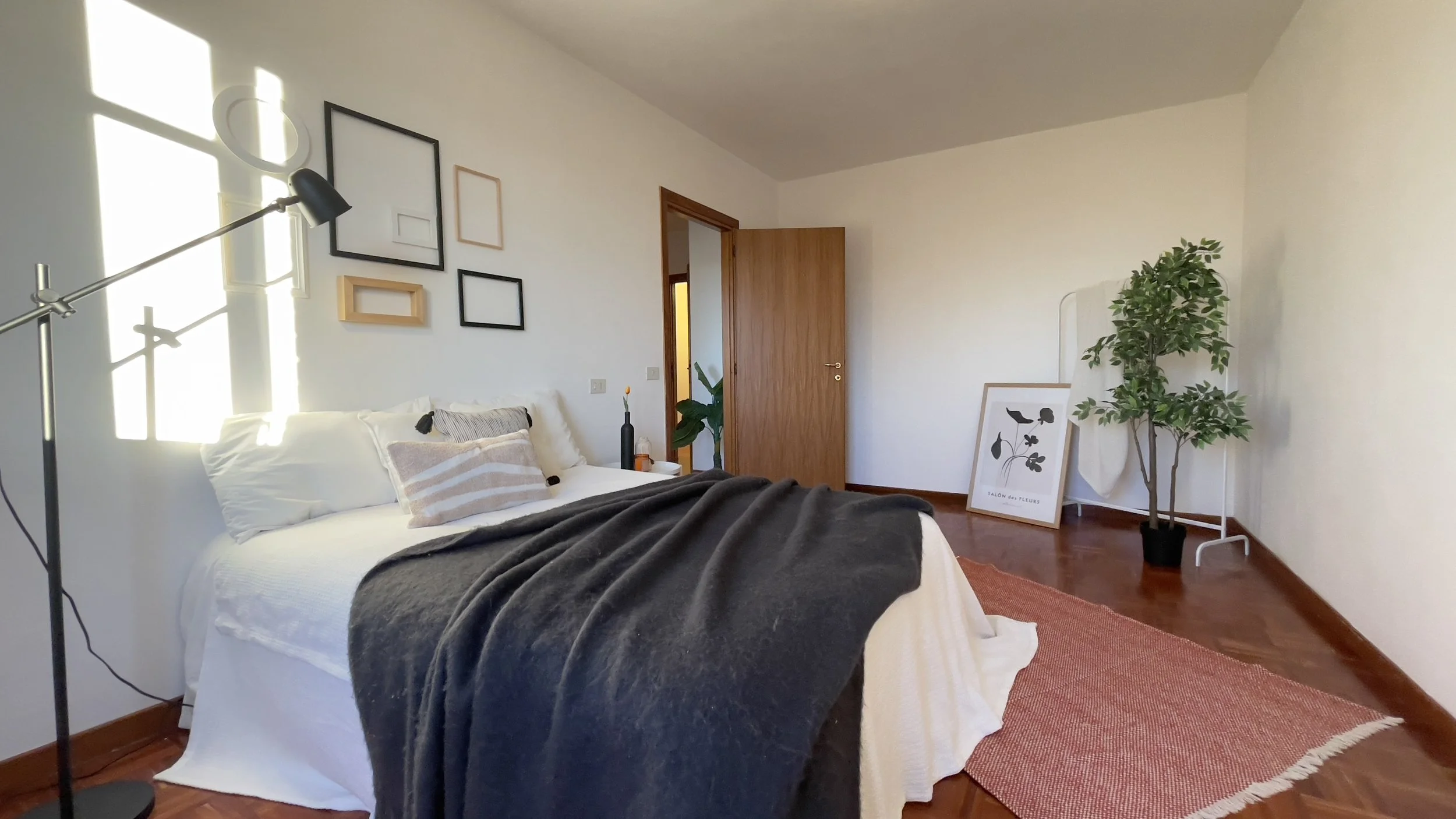 Camera da letto minimalista con letto bianco, coperte grigie, lampada da lettura, cornici decorative vuote sul muro, pianta in vaso e tappeto rosso.