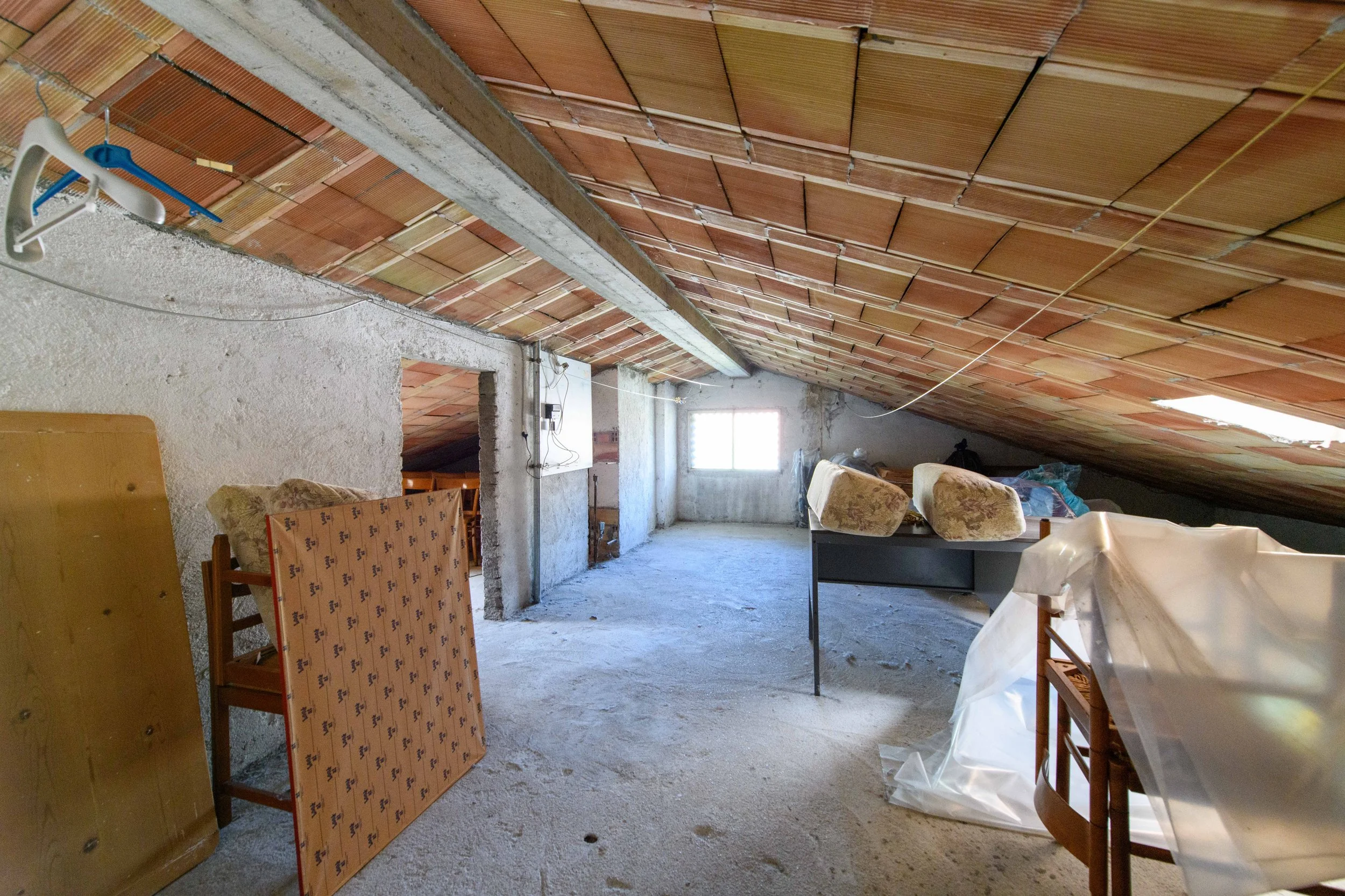 Soffitta disordinata con tetto spiovente, oggetti coperti da teli, materassi appoggiati, sedie e una finestra sul fondo.