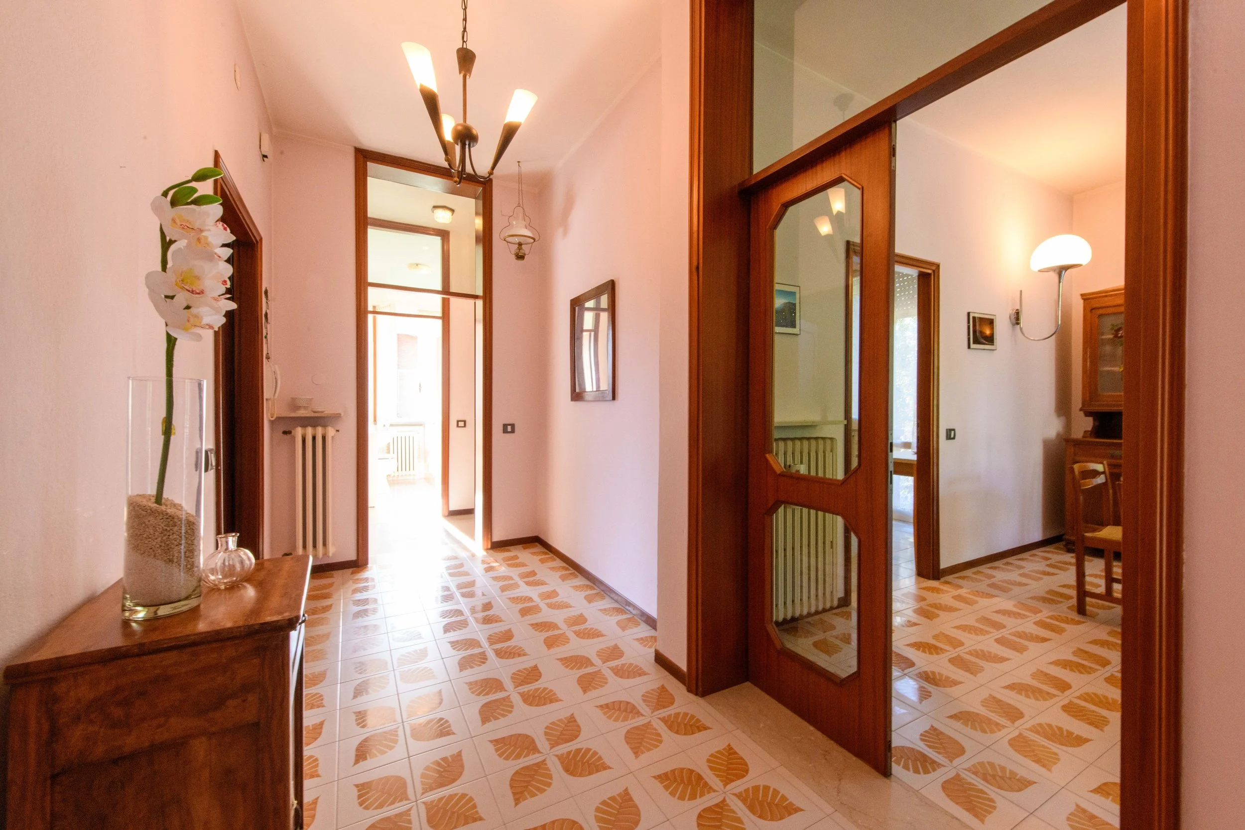 Interno di un appartamento con corridoio, piastrelle a motivi geometrici, porta in legno con vetri, lampadario moderno e specchio sulla parete.