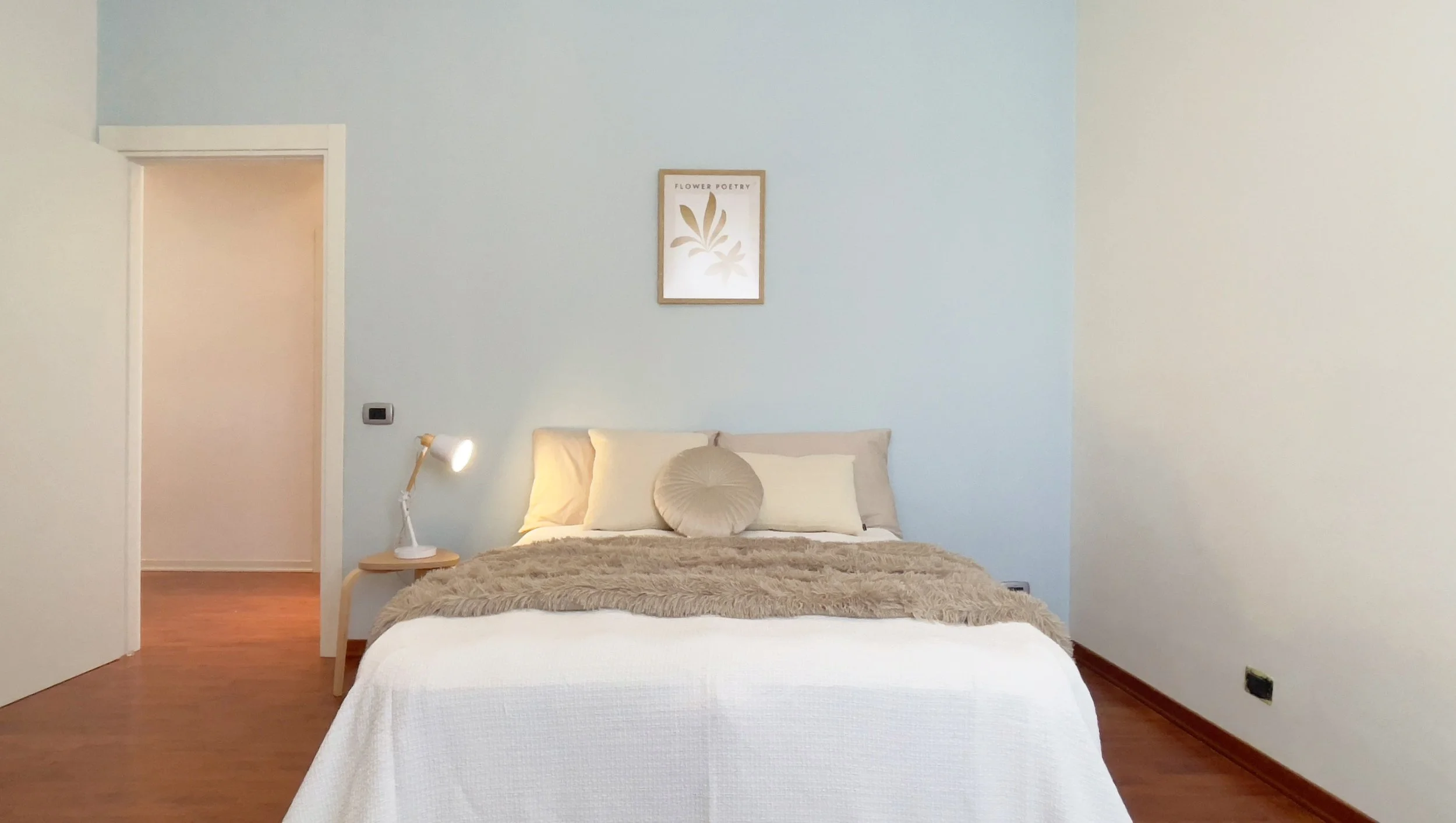 Camera da letto con letto matrimoniale, cuscini bianchi e beige, coperta pelosa color crema, lampada da tavolo e quadro decorativo sulla parete.