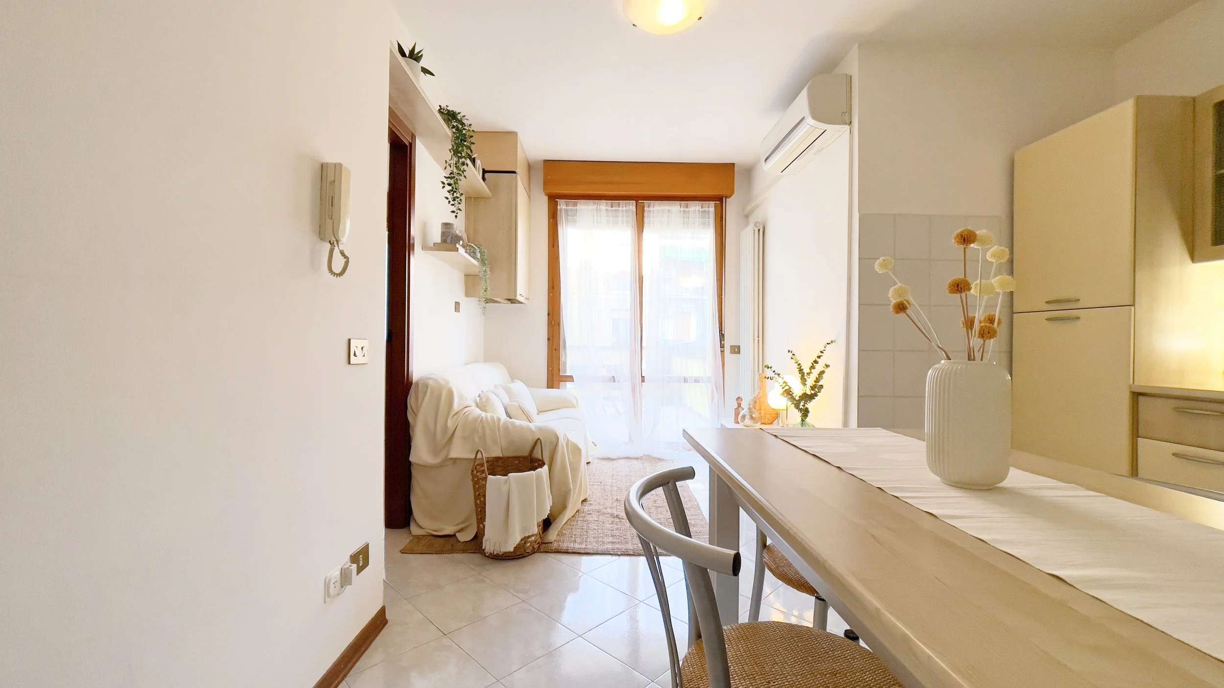 Interno di un appartamento luminoso con soggiorno e zona pranzo, arredato in stile moderno. Il soggiorno ha un divano bianco coperto con un telo, una mensola con piante e decorazioni, e una grande finestra con tenda trasparente. Nella zona pranzo c'è