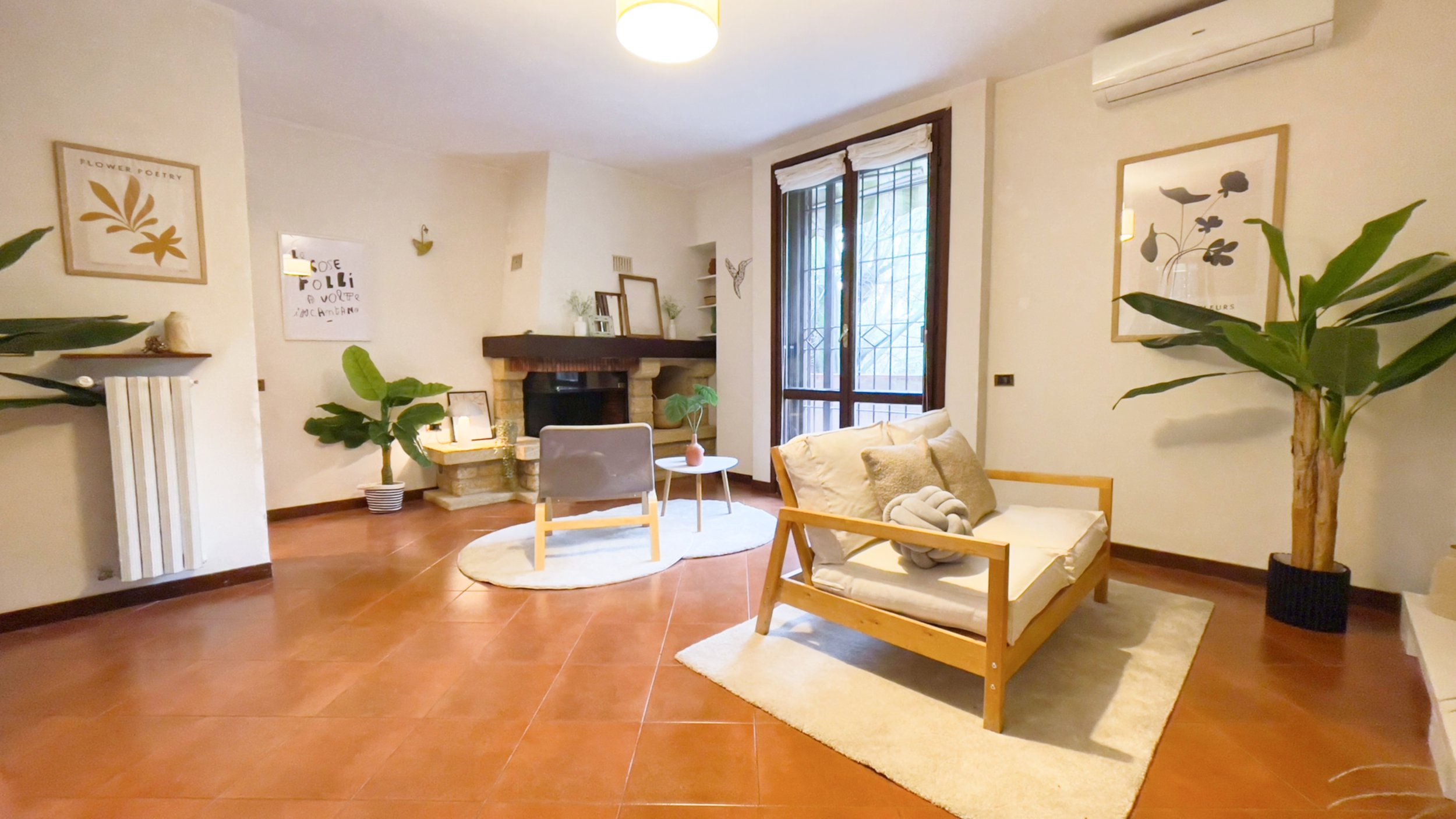 Stanza luminosa e accogliente con un divano in legno chiaro e cuscini chiari, un tavolino, una poltrona e piante verdi. C'è un camino in un angolo e pareti con decorazioni artistiche. Ampia finestra con persiane e un'unità di condizionamento sopra.