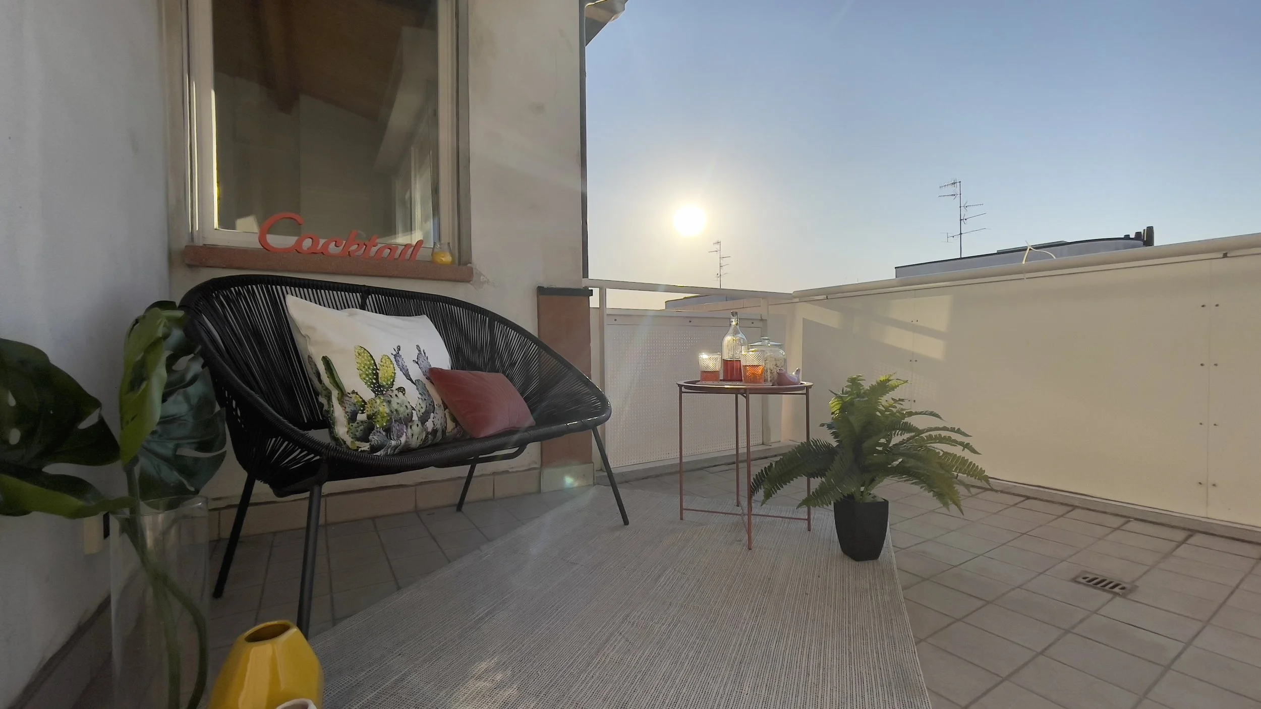 Terrazza arredata con sedia nera, cuscini decorativi, tavolino con drink e decorazioni, pianta verde e scritta "Cocktail" sul davanzale. Sole al tramonto sullo sfondo.