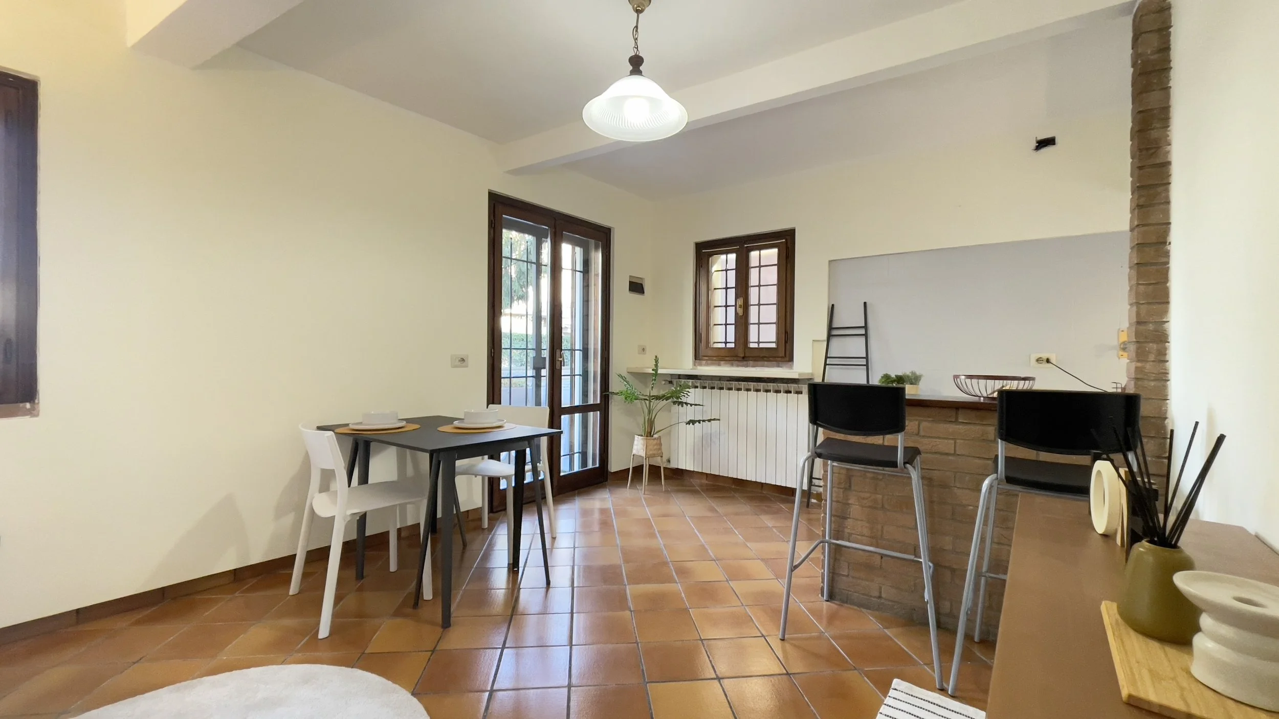 Sala da pranzo con tavolo e sedie, pavimento in piastrelle color terracotta, porta finestra e finestra, bar con sgabelli, decorazioni minimaliste.