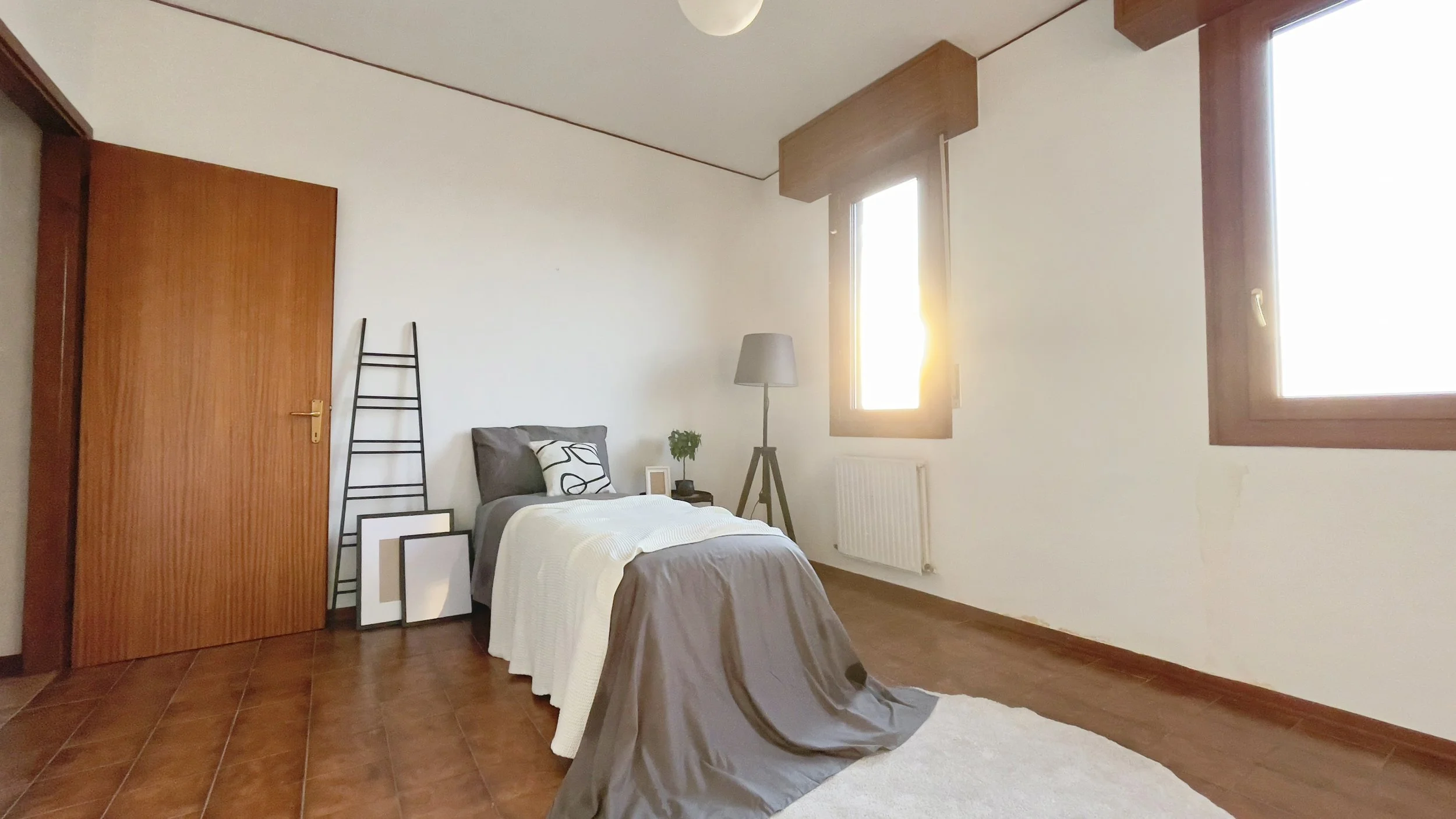 Camera da letto moderna con letto singolo, copriletto grigio e bianco, lampada da terra, finestra con luce naturale, pavimento in legno e scala decorativa.