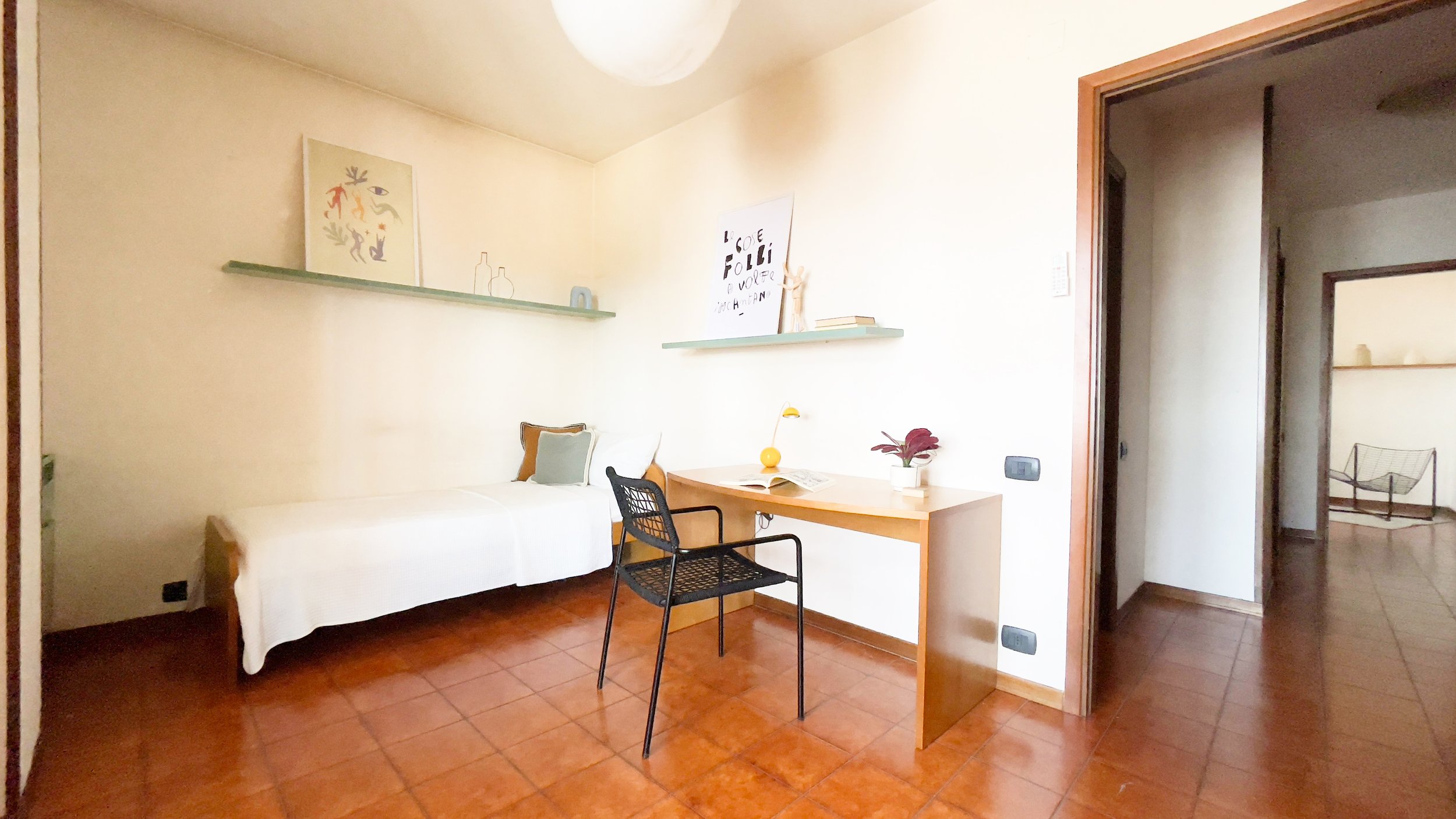Stanza minimalista con letto singolo, scrivania e sedia, decorata con mensole e quadri su pareti chiare.