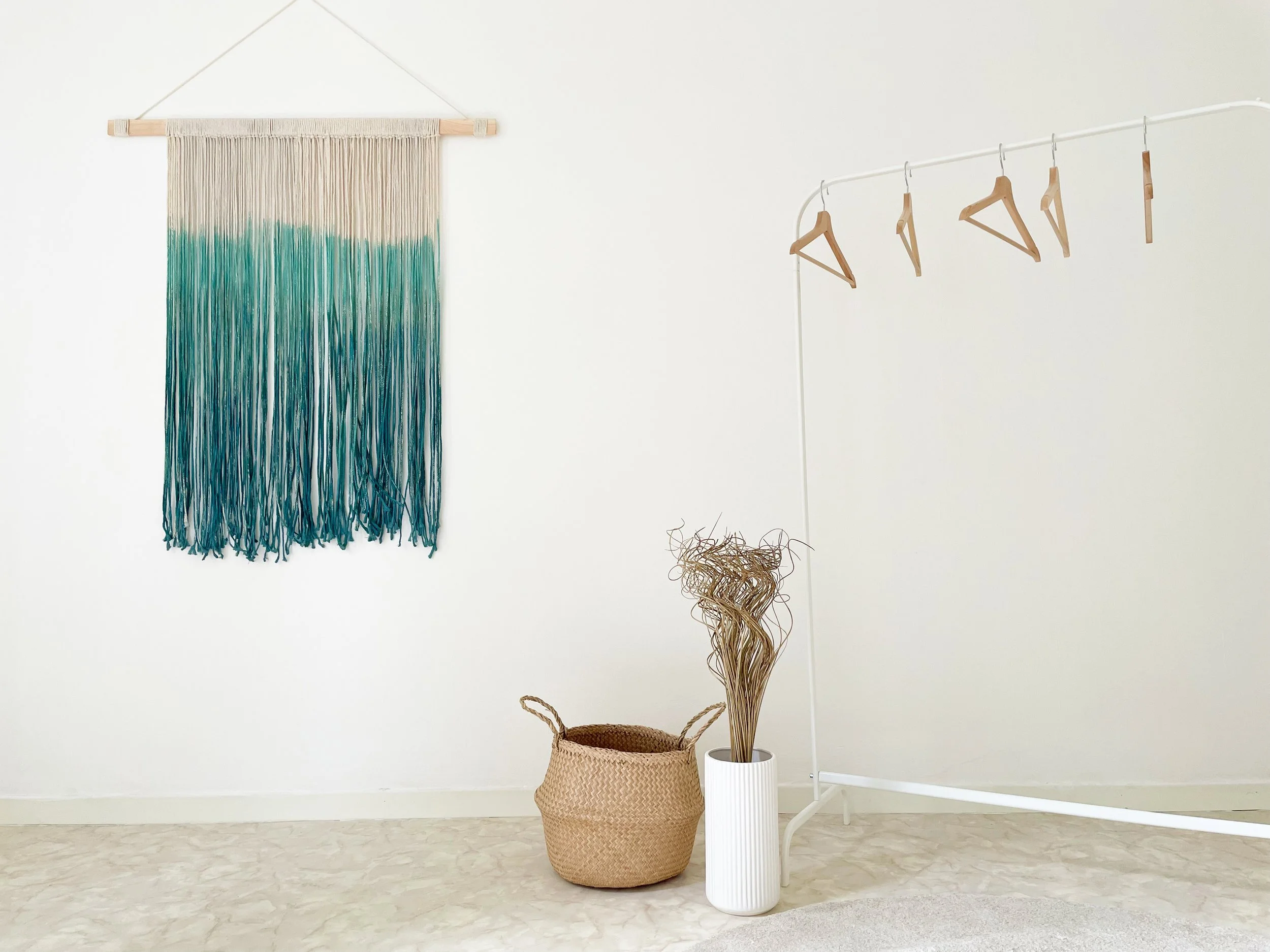 Interno minimalista con una decorazione da parete macramè blu e bianca, un cesto di vimini, due vasi bianchi, e appendiabiti vuoti su un supporto bianco.