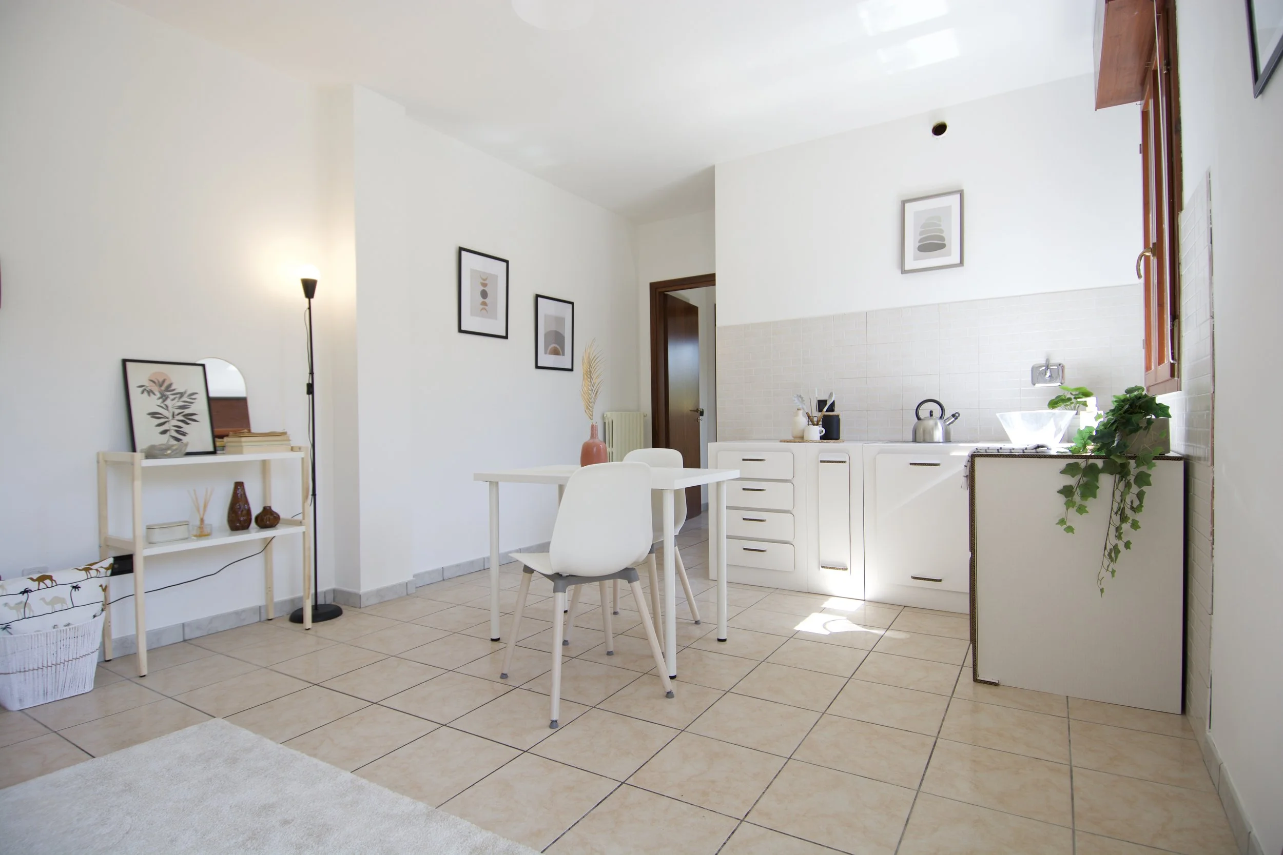 Soggiorno moderno con cucina a vista. Tavolo bianco, due sedie, pareti bianche con quadri astratti. Cucina con armadietti bianchi, pianta in vaso e scaldavivande.