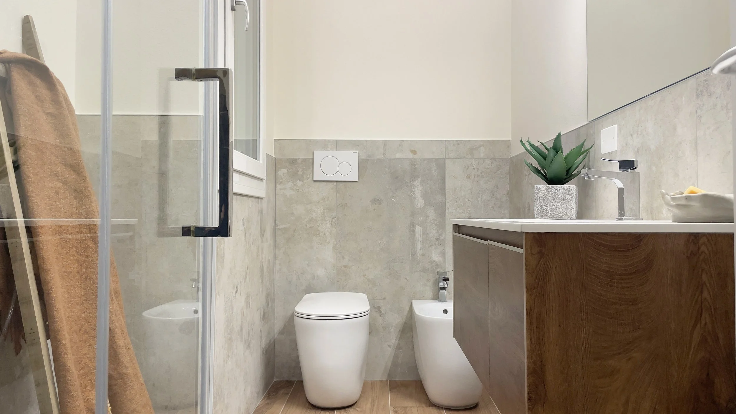 Bagno moderno con servizi igienici, pareti in piastrelle grigie e pavimento in legno. Lavabo con rubinetto cromato e pianta decorativa, asciugamano arancione appeso.